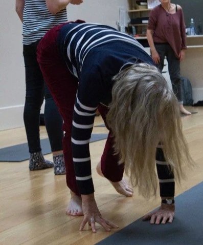 Person in a striped shirt and red pants bending forward, with others standing nearby, practicing yoga indoors.