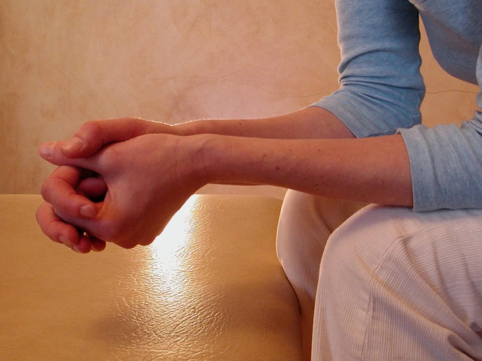Person sitting with hands clasped, wearing a light blue long sleeve shirt and beige pants, on a tan surface with a plain background.