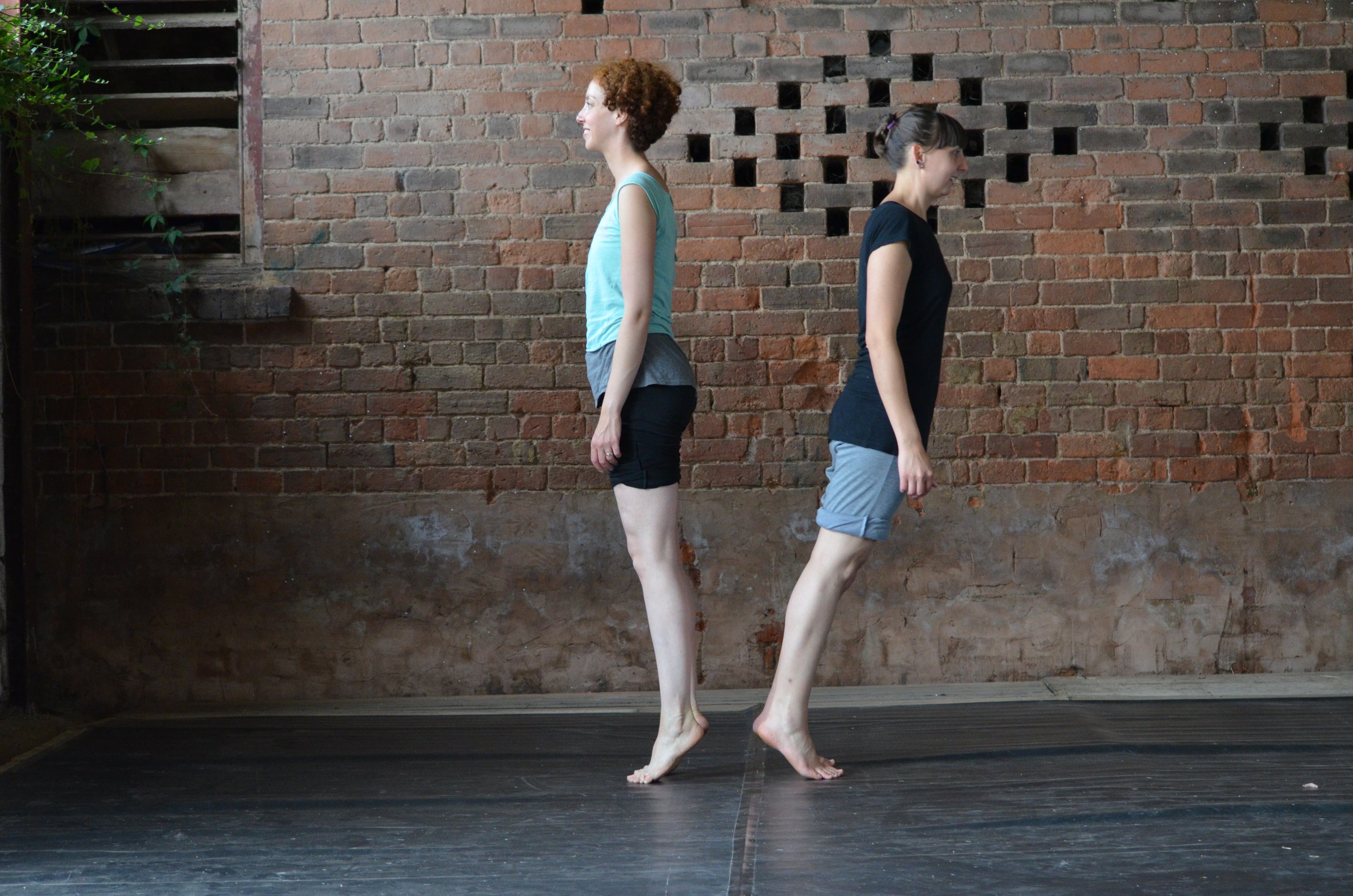 Two women are standing back-to-back on a wooden floor, with their heels touching and toes pointed outward, in an outdoor setting with an exposed brick wall behind them.