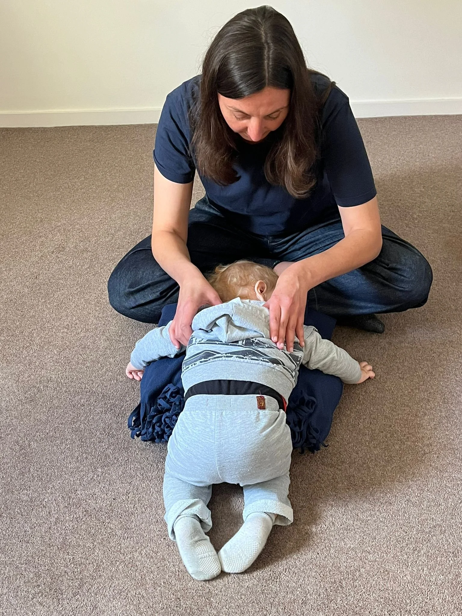 A woman practicing child first aid on a small child lying face down on a carpeted floor.