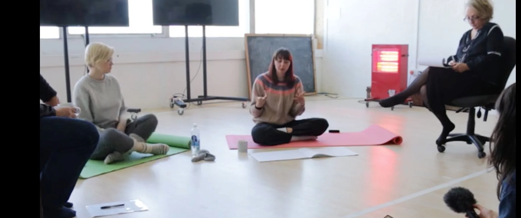 Group of people sitting on the floor in a circle, participating in a discussion or workshop, with a woman in a black outfit sitting on an office chair, and other items like water bottle, papers, and mats on the floor.