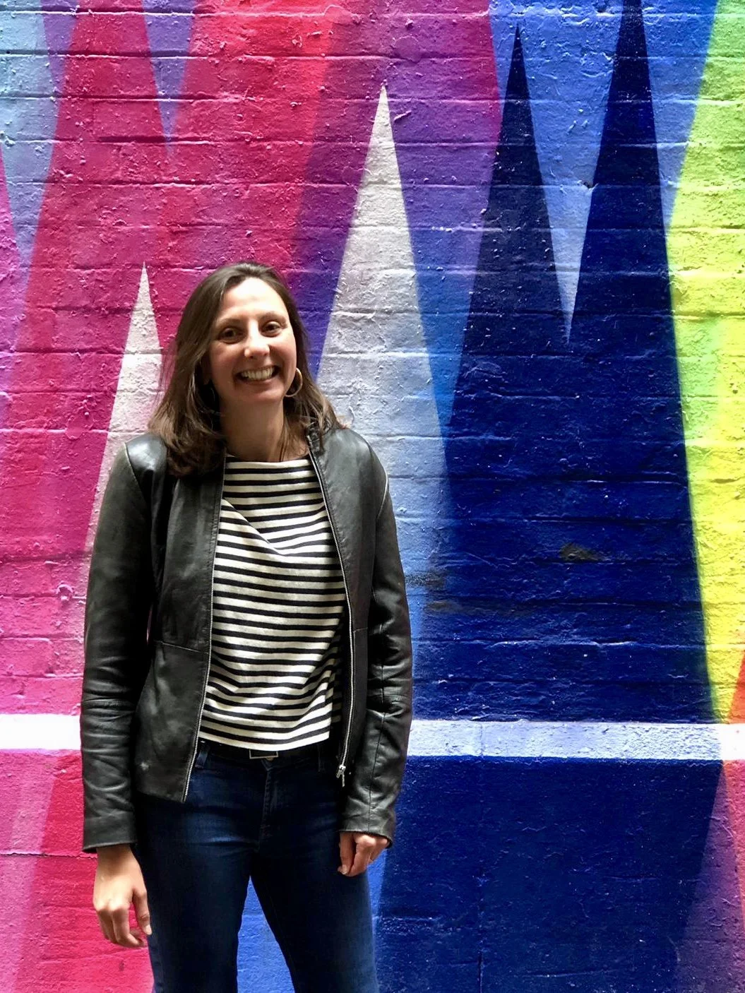 A smiling woman with shoulder-length brown hair, wearing a black leather jacket and a black-and-white striped shirt, standing in front of a colorful graffiti wall.