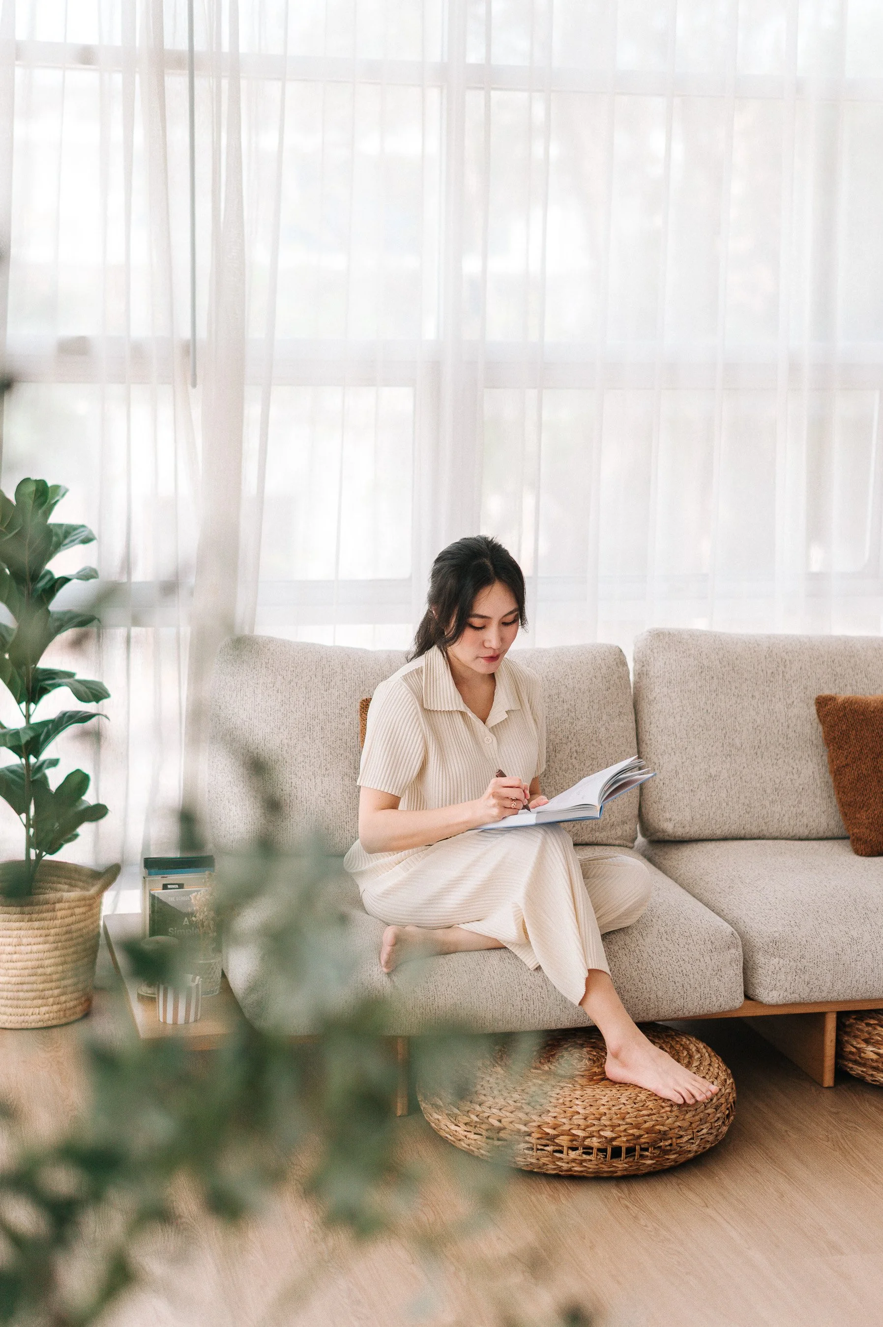 A woman sitting on a beige sofa in a bright living room, writing in a notebook.