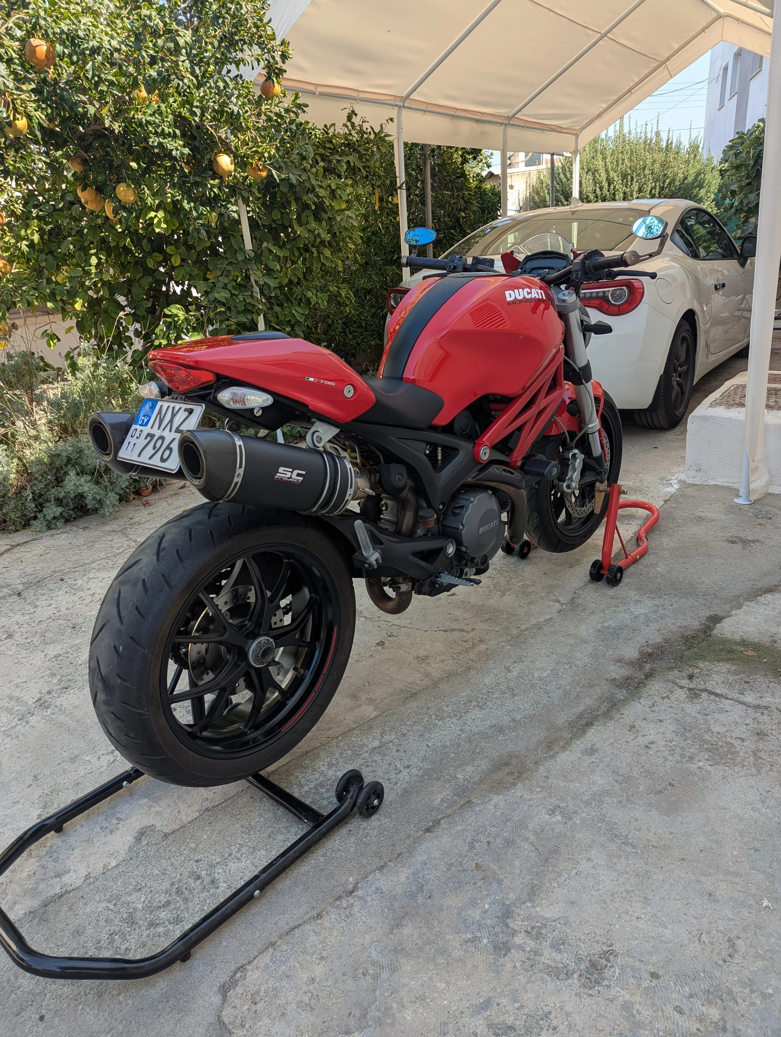 A red Ducati motorcycle on a paddock stand next to a white sports car under a canopy, with greenery and orange trees in the background.