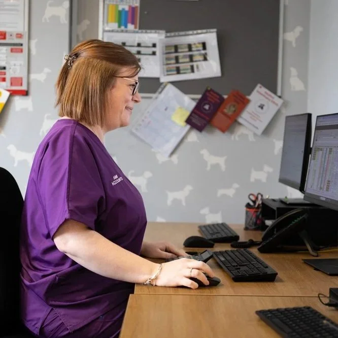 A woman with red hair and glasses working at a desk with a computer in an office environment. She is wearing purple scrubs and is smiling at her screen.