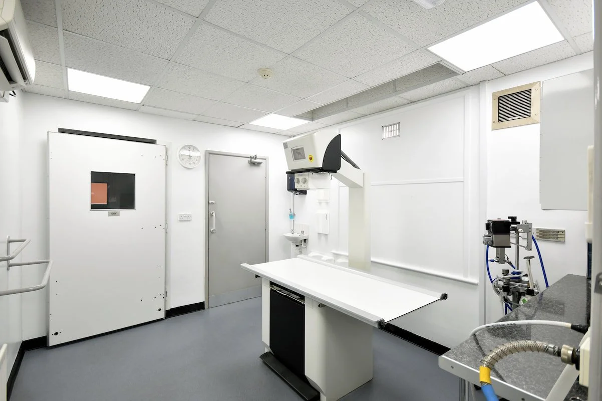 A clean, sterile medical imaging room in Godiva Vet Clinic with white walls and ceiling, featuring X-ray equipment, a patient table, and various medical devices.
