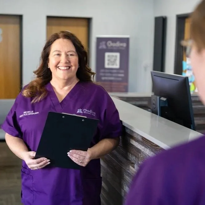 Smiling woman wearing a purple uniform standing at a reception desk, holding a clipboard, with a person partially visible in the foreground.