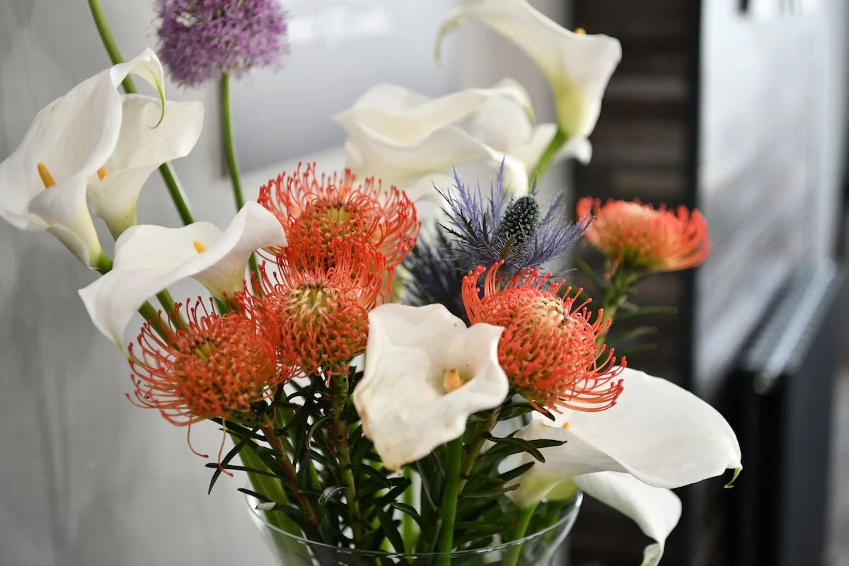 A bouquet of white calla lilies, red protea flowers, and other colorful flowers in a glass vase.