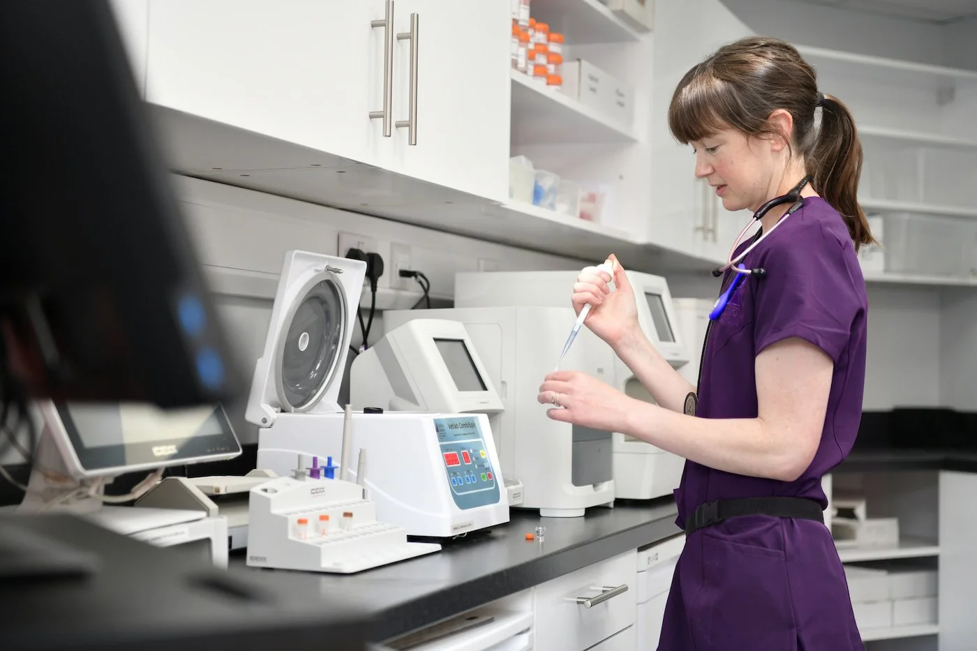 A young female healthcare professional in purple scrubs and a stethoscope around her neck working in a laboratory, handling a test tube with laboratory equipment on the counter.