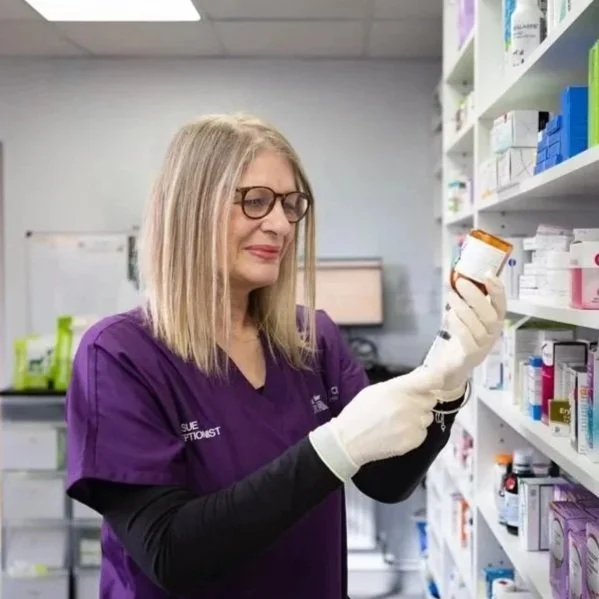 A woman in purple pharmacy scrubs and glasses, wearing white gloves, holding a medication bottle in a pharmacy aisle.