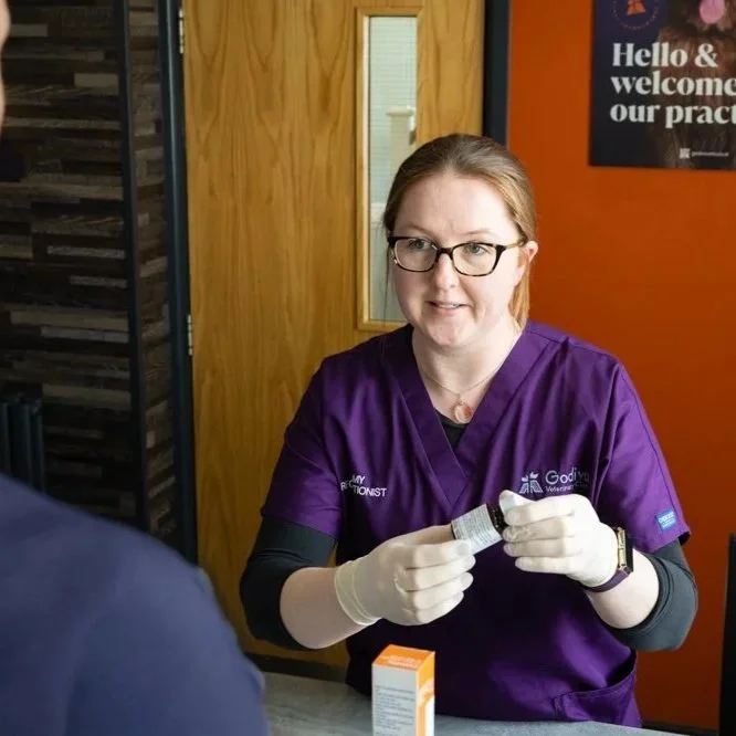 A woman wearing purple scrubs and glasses is preparing a medical injection, sitting at a desk with a small orange and white box in front of her.