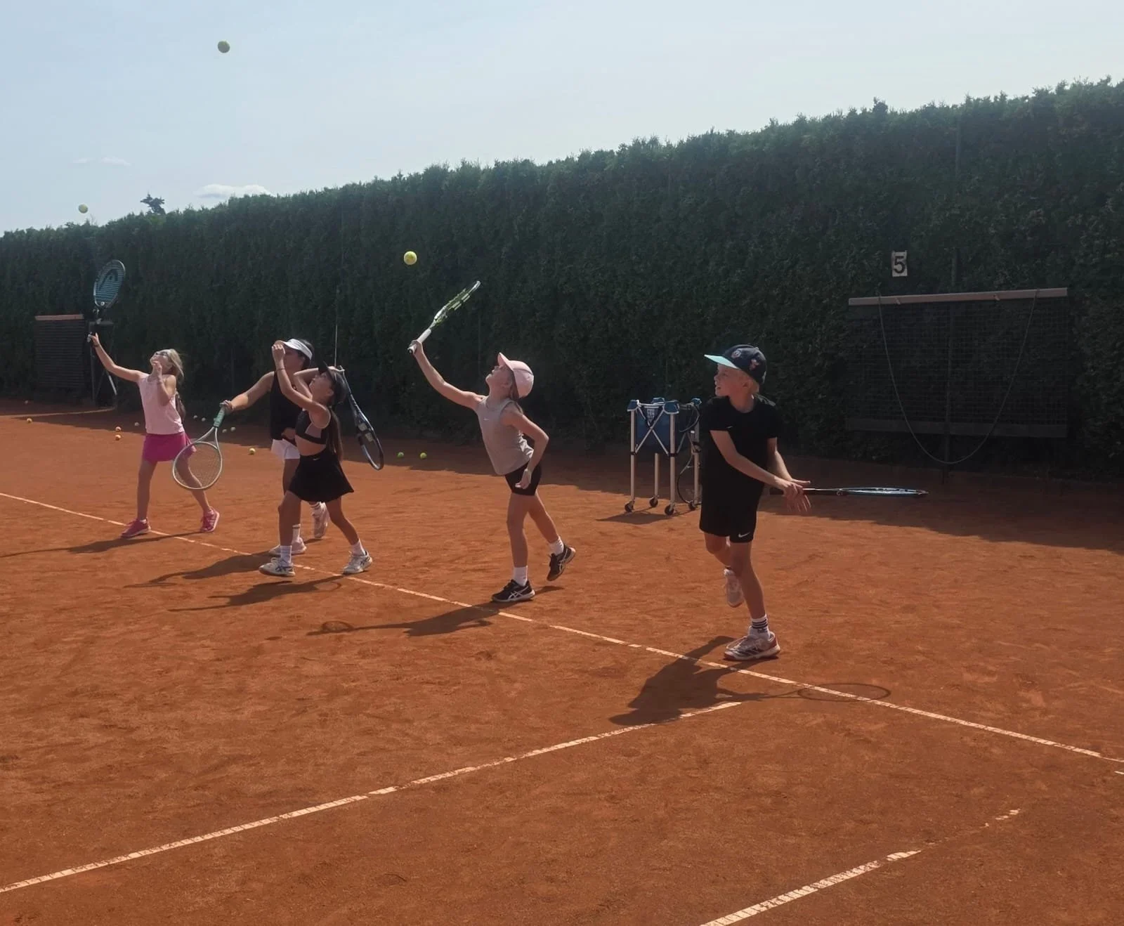 Children practicing tennis on a clay court during daytime, with five kids holding tennis rackets and hitting tennis balls, three girls and two boys, with a tall hedge in the background.