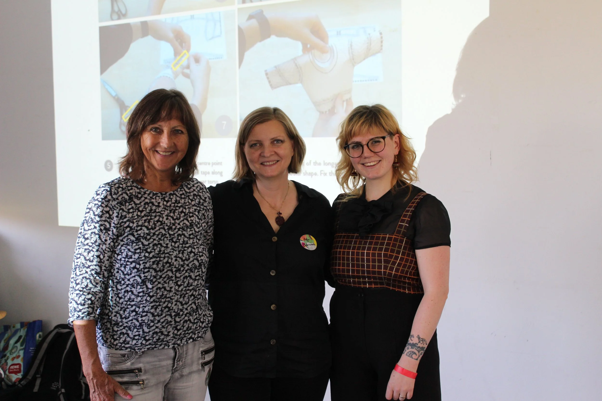 Three women standing together indoors in front of a projected presentation, smiling at the camera.