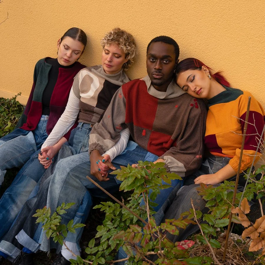 Four young adults sitting against a yellow wall with their eyes closed, holding hands, and leaning on each other, surrounded by some plants.
