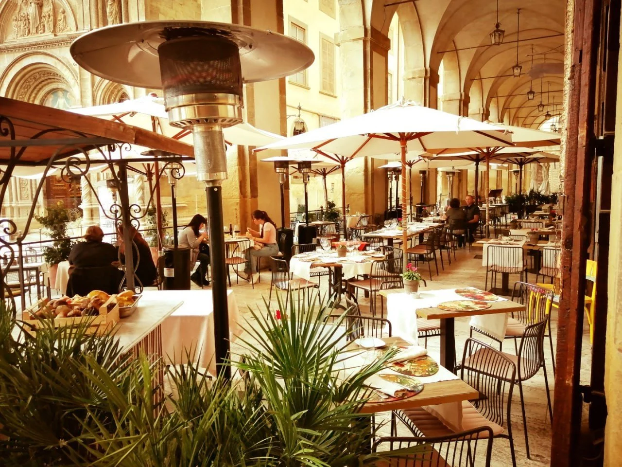 Outdoor cafe or restaurant with white umbrellas, tables set for dining, and people sitting at tables inside a historic building with arches and ornate architecture.