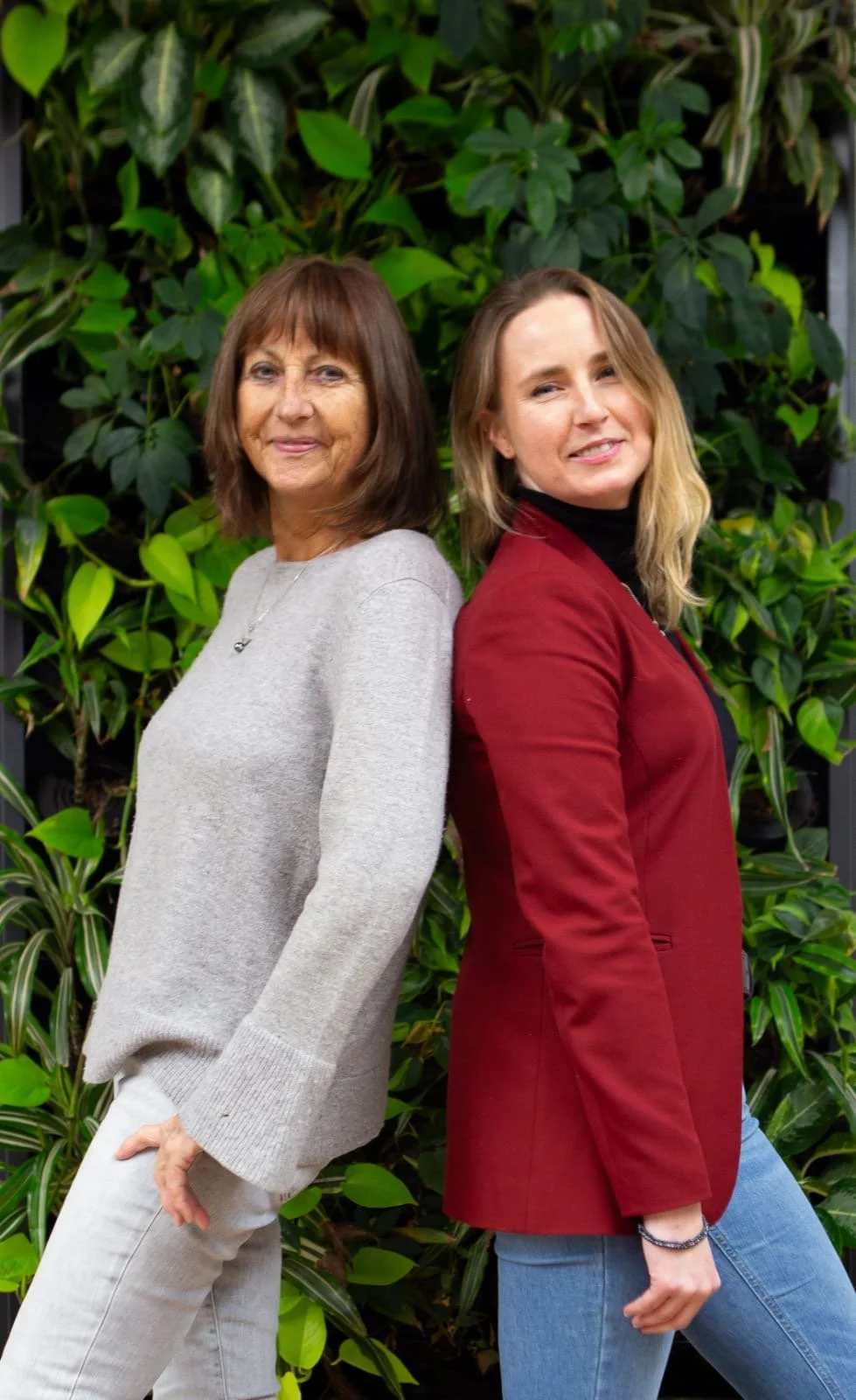 Two women are standing back to back outdoors in front of lush green plants. The older woman on the left has shoulder-length brown hair and is wearing a light gray sweater and white jeans, smiling gently. The younger woman on the right has shoulder-length blonde hair and is wearing a black turtleneck, a red blazer, and blue jeans, also smiling.