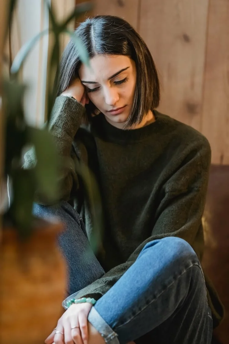 A young woman with shoulder-length dark hair, wearing a dark sweater and jeans, sitting with her eyes closed and resting her head on her hand in a contemplative pose in a cozy, wooden-paneled room.
