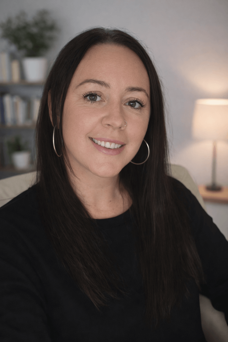 A smiling woman with long dark hair, wearing hoop earrings and a nose ring, sitting in a cozy room with a bookshelf and a lit lamp in the background.