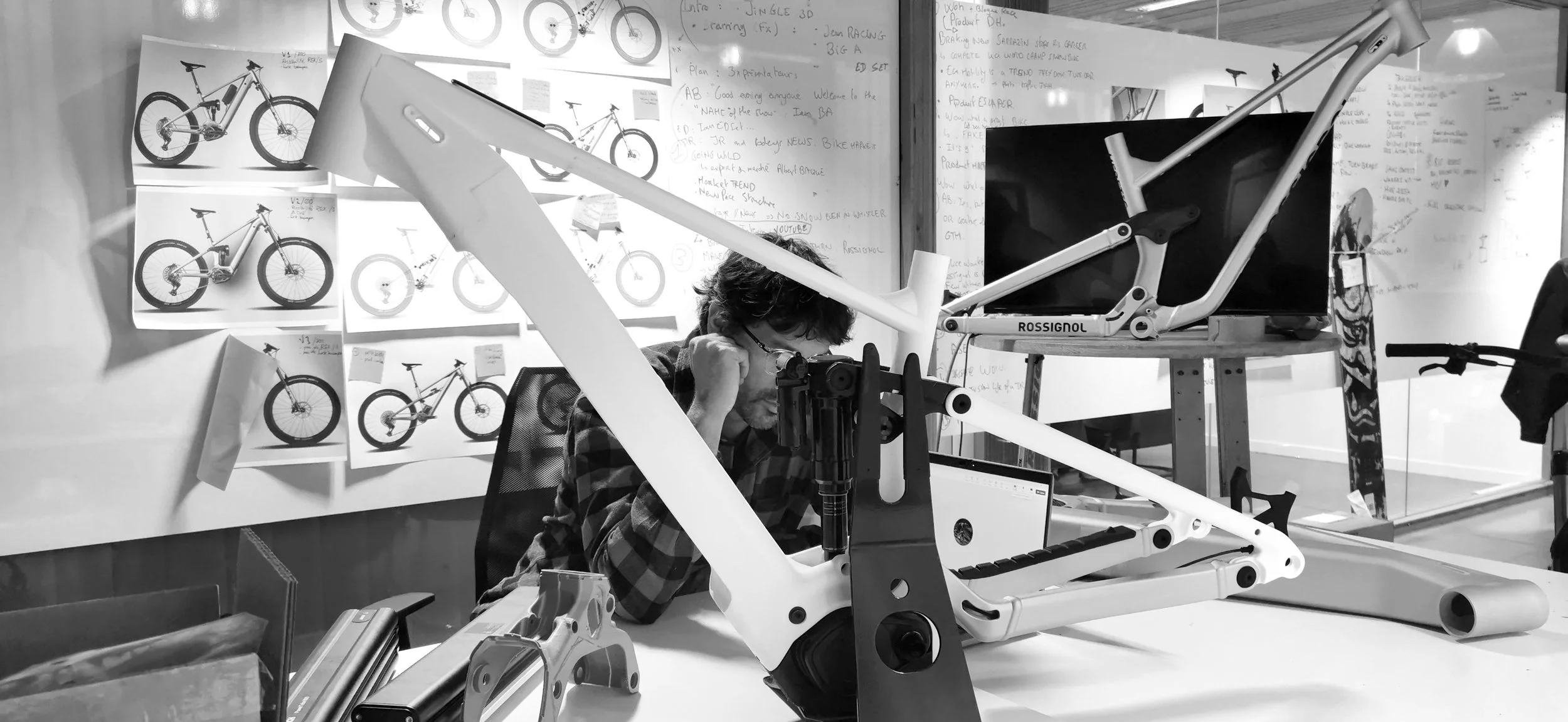 Un homme travaillant sur un cadre de vélo dans un atelier, avec des plans de vélos affichés au mur en arrière-plan.