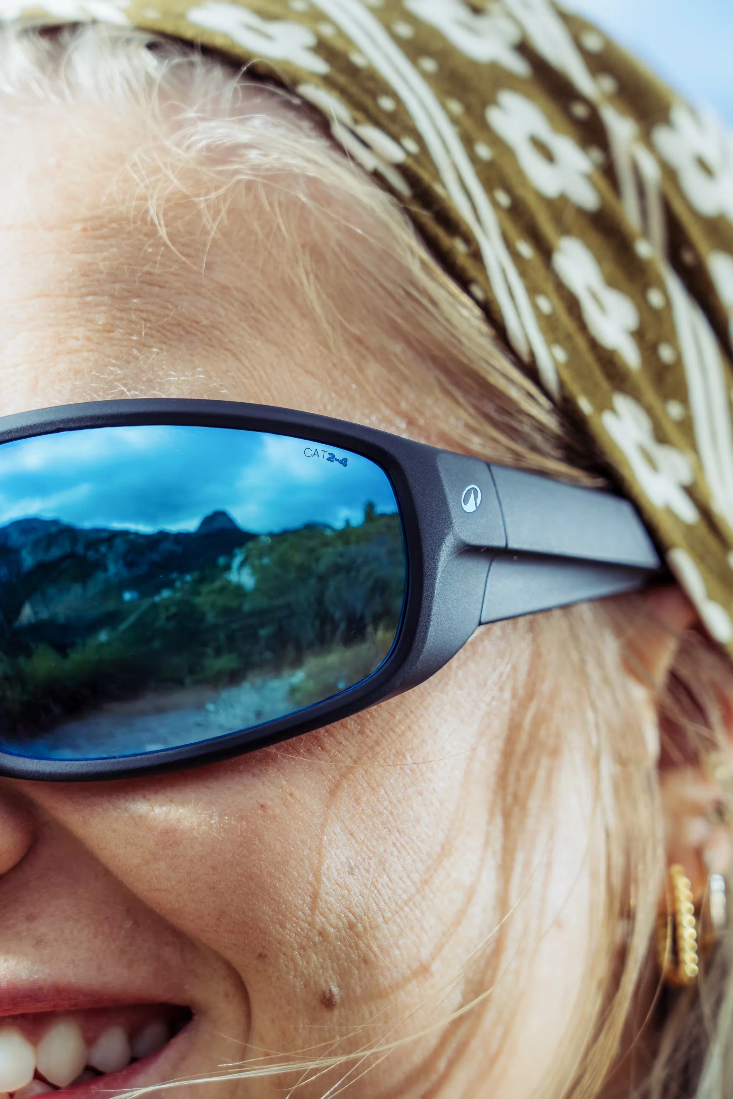 Une femme portant des lunettes de soleil réfléchissant la nature, souriant et proche de la caméra, avec des cheveux blonds, des taches de rousseur et des bijoux dorés.