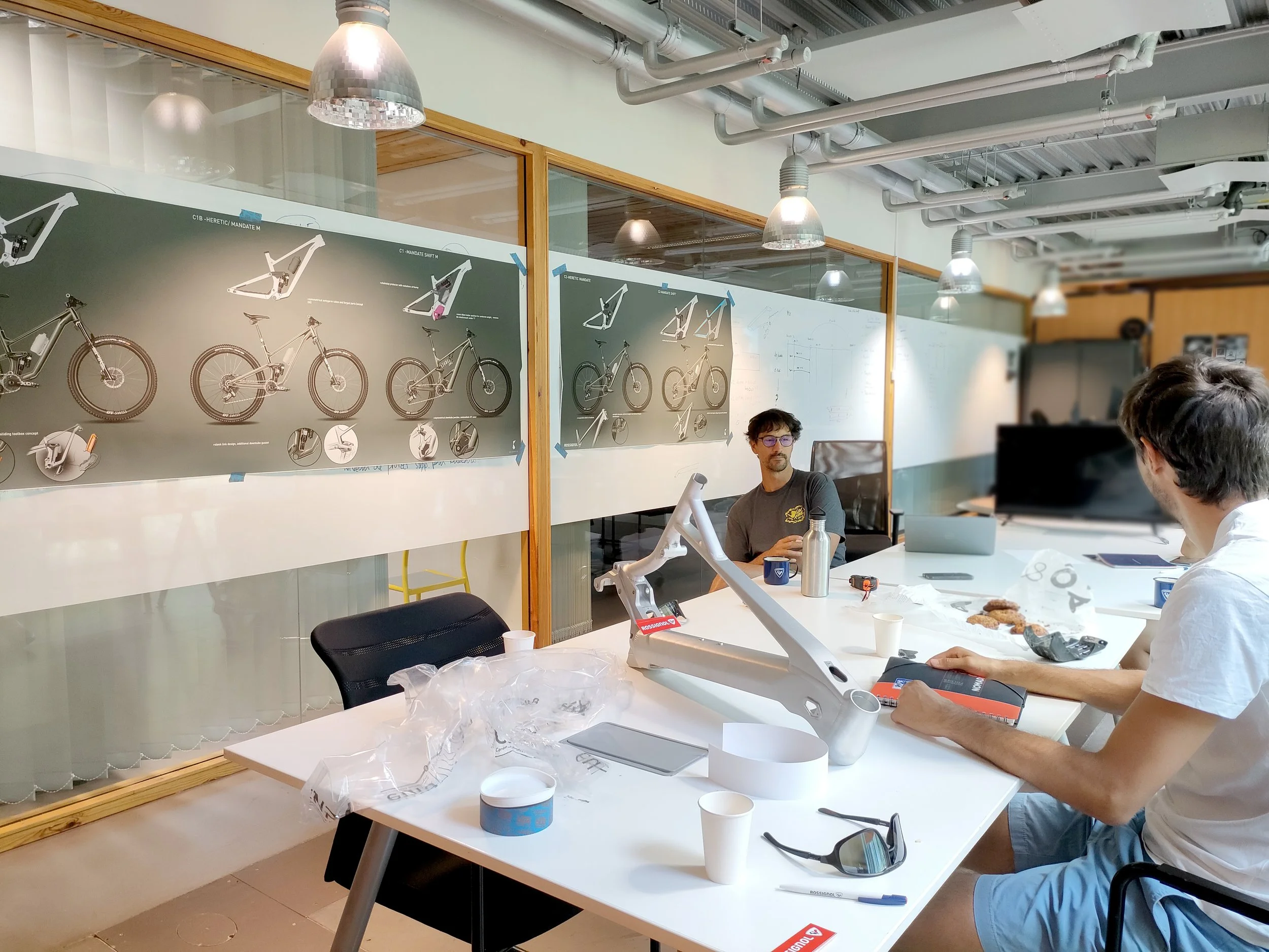 Une salle de réunion ou d'atelier avec plusieurs personnes. Sur le mur, affiches de prototypes de vélos ou d'équipements liés à la bicyclette, avec des dessins techniques. La pièce est moderne, avec des luminaires suspendus et des conduits aériens au plafond.