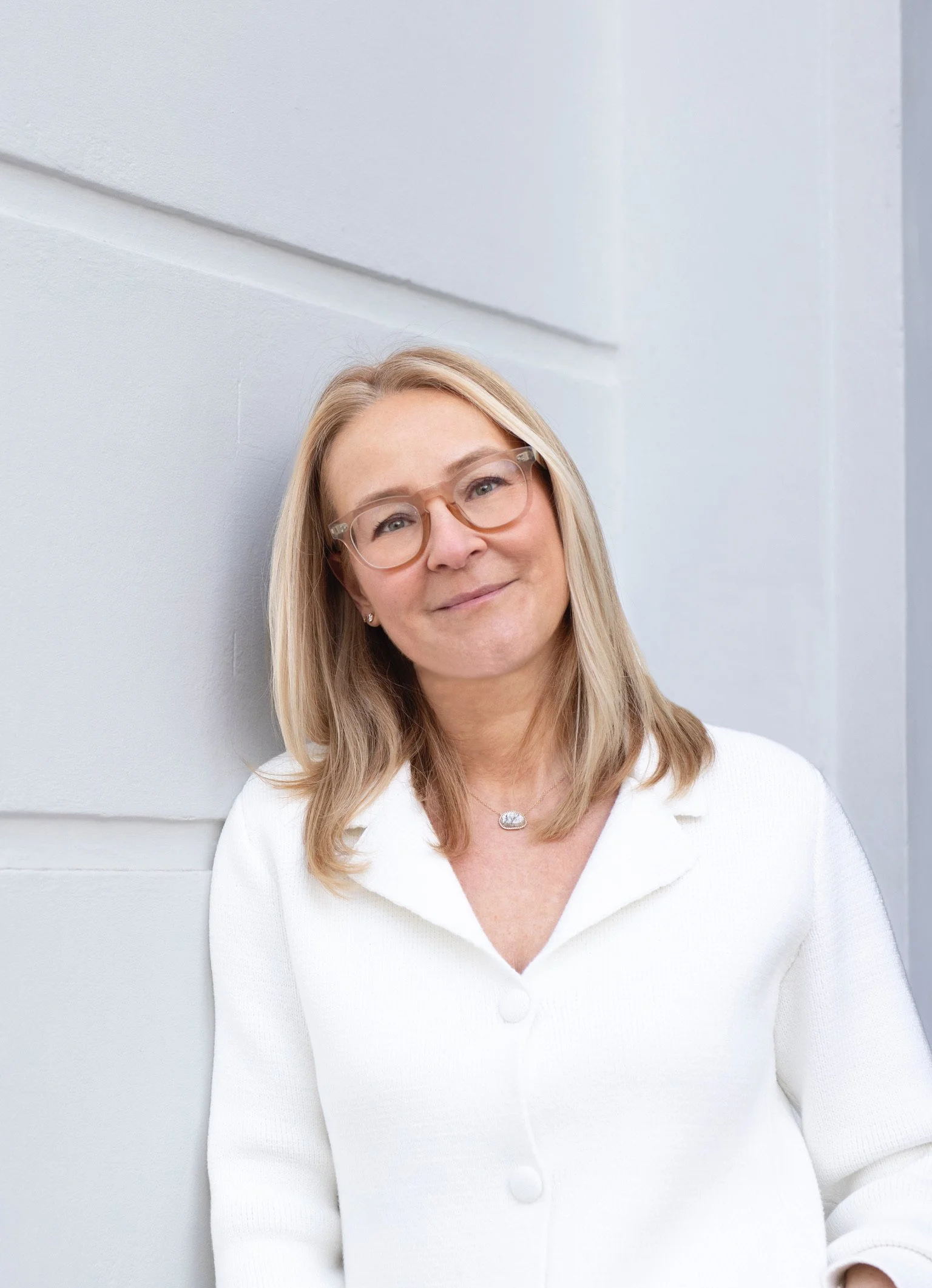 A woman with blonde hair and glasses wearing a white shirt, smiling and leaning against a white wall.