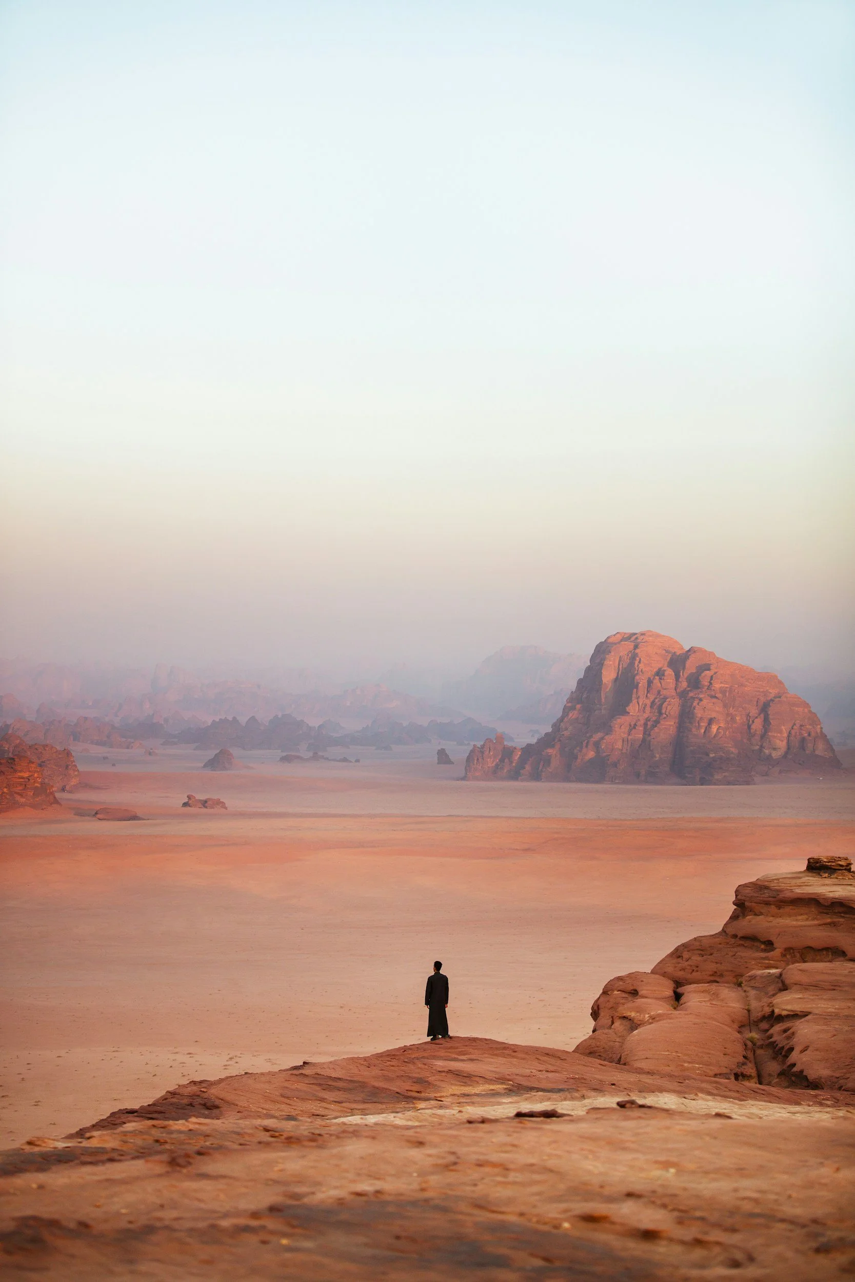 Desert landscape with large rocky mountains, a palm tree, and a person riding a camel under a clear blue sky.