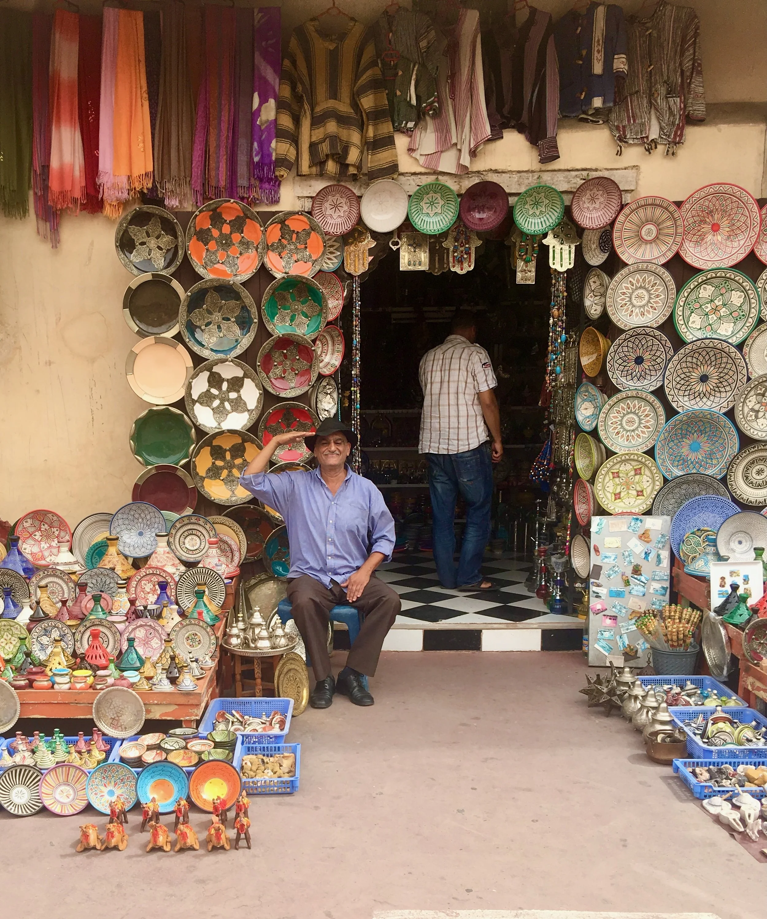 A man sitting outside a shop selling colorful traditional hand-painted bowls, plates, and souvenirs. The shop has items displayed on walls, tables, and in baskets.