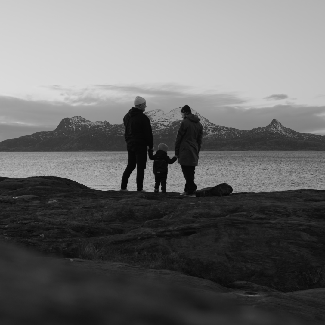 En familie står ved vannet og ser mot fjellene i bakgrunnen.