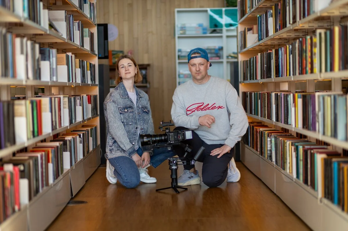 To personer kneler mellom lange bokhyller i et moderne bibliotek og poserer ved siden av et profesjonelt filmkamera.