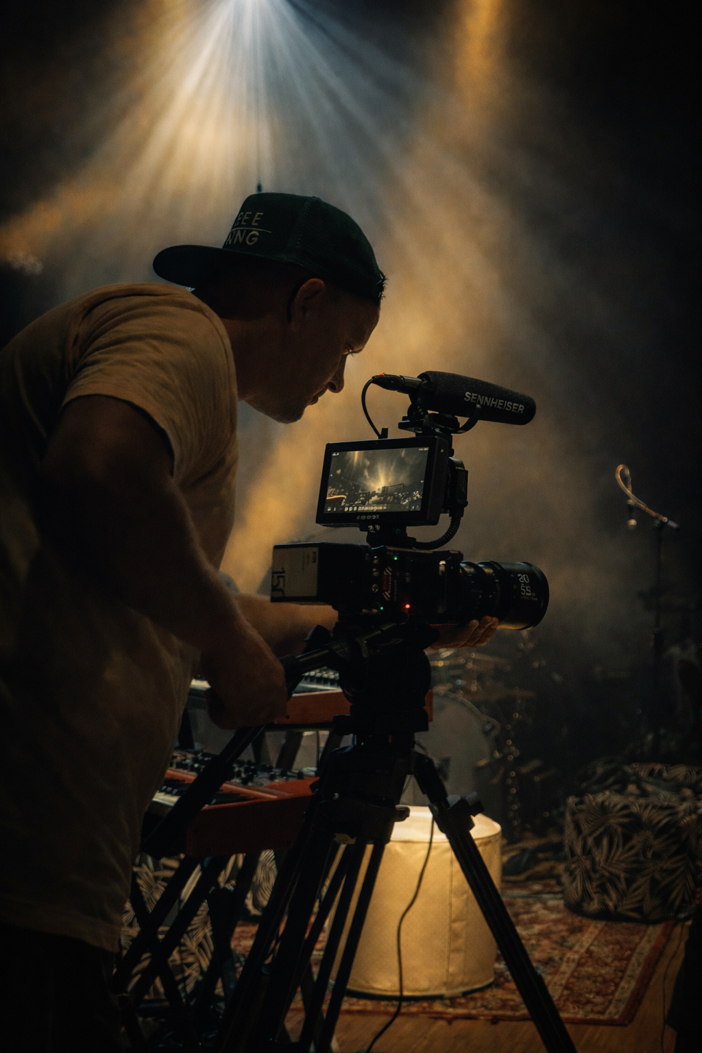 En kameramann i baseballcaps justerer et filmkamera montert på et stativ mens han filmer en livekonsert.
