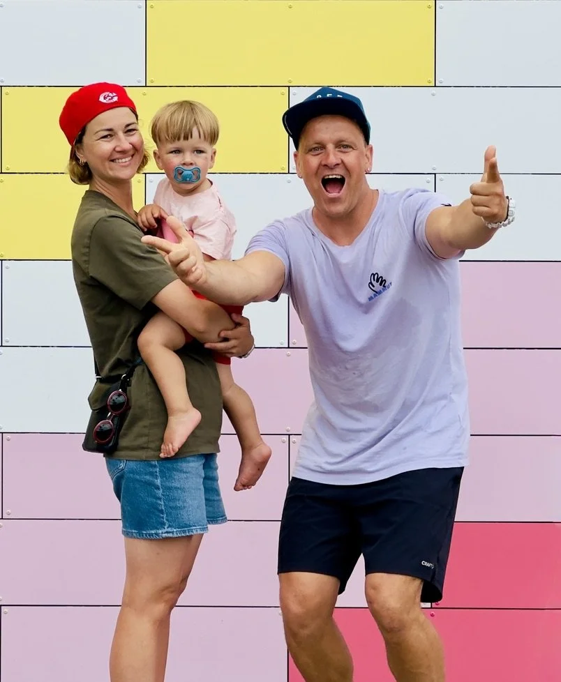 En familie poserer foran en fargerik vegg: en kvinne holder et lite barn med smokk, mens en mann ved siden av smiler.