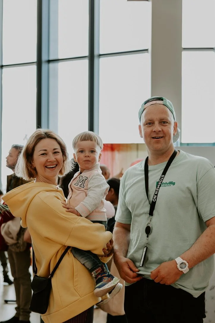En familie smiler innendørs: en kvinne holder et barn, mens en mann ser mot kameraet. Frivillige på internasjonal barnedag i biblioteket.