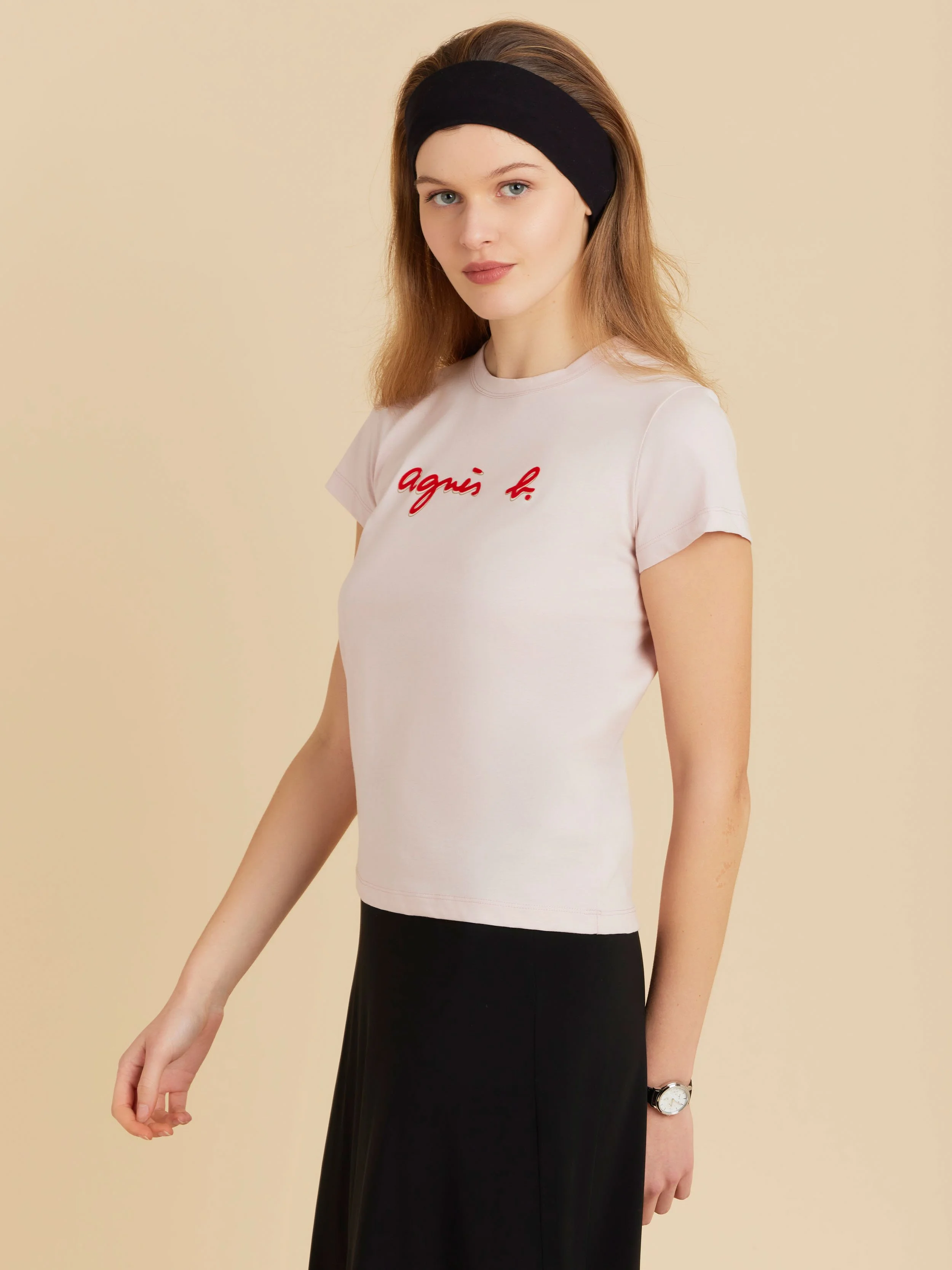 Jeune femme portant une t-shirt blanc avec le nom 'agnès b.' en rouge, une montre noire, un bandeau noir et un pantalon noir, contre un fond beige clair.
