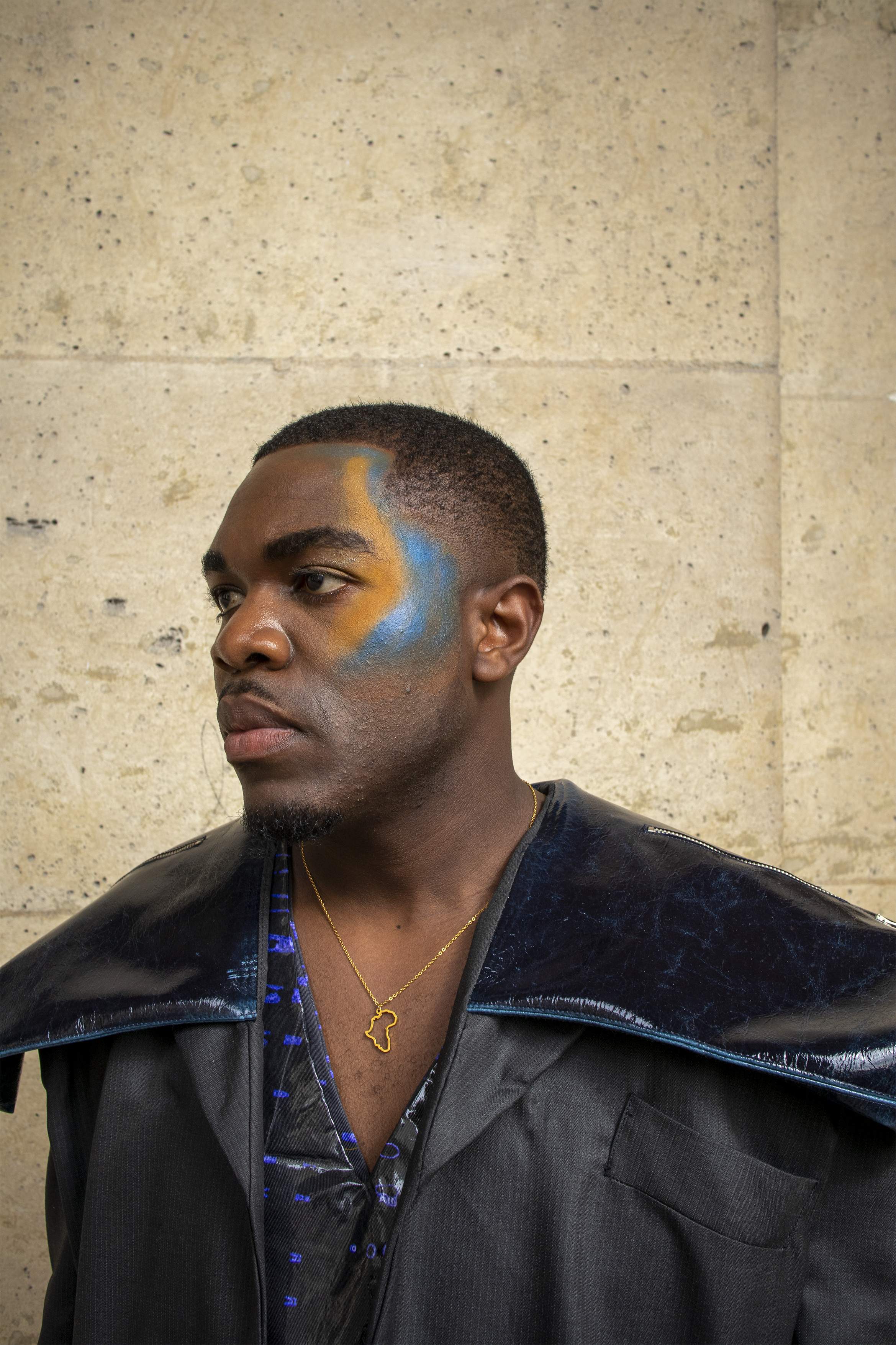 Portrait d'un homme avec un tatouage bleu et jaune sur le visage, portant une veste noire avec un col en plastique noir brillant, et portant un collier en forme de l'Afrique