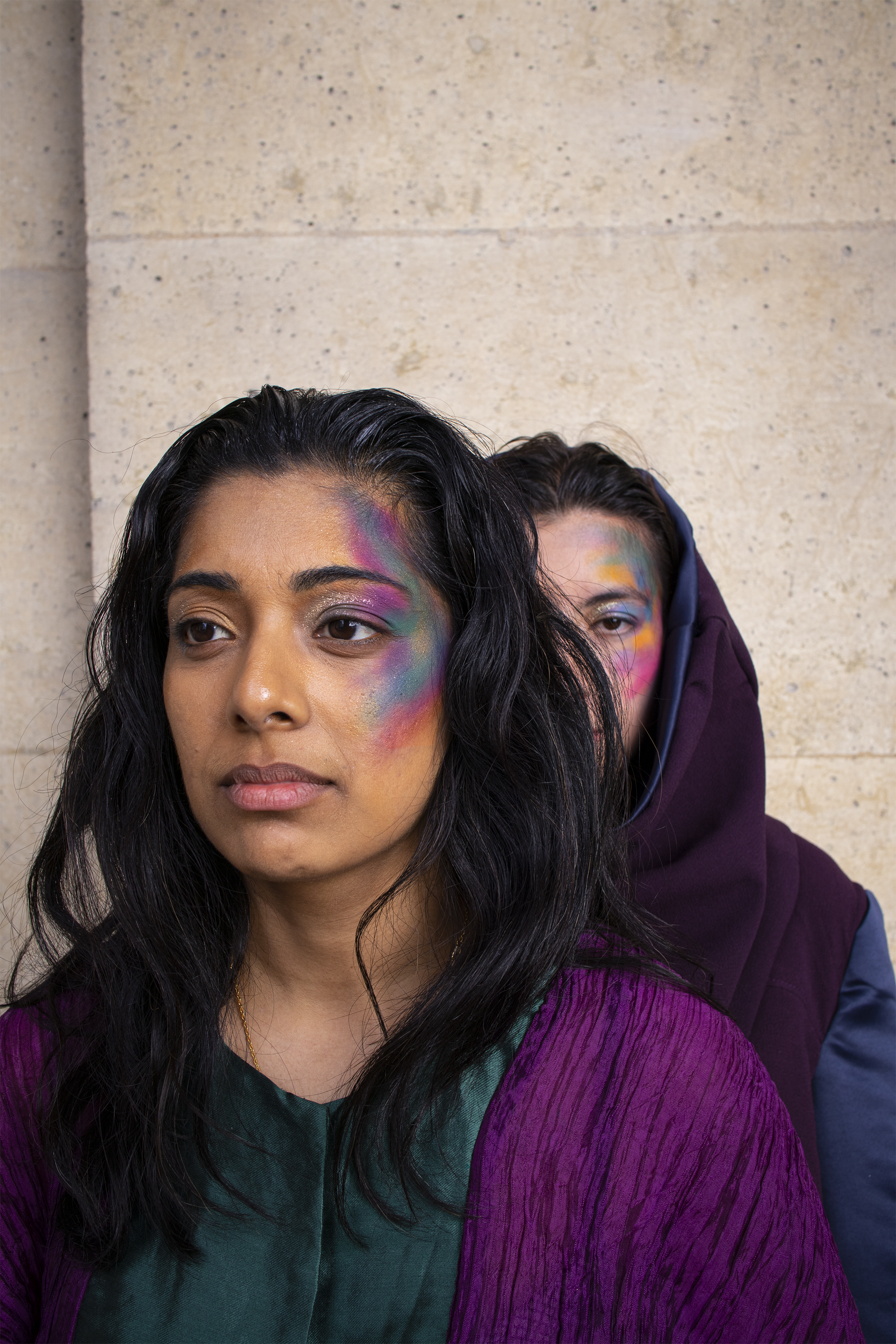 Deux femmes debout contre un mur en pierre, avec des maquillages colorés sur le visage, l'une devant et l'autre à l'arrière, partiellement cachée.