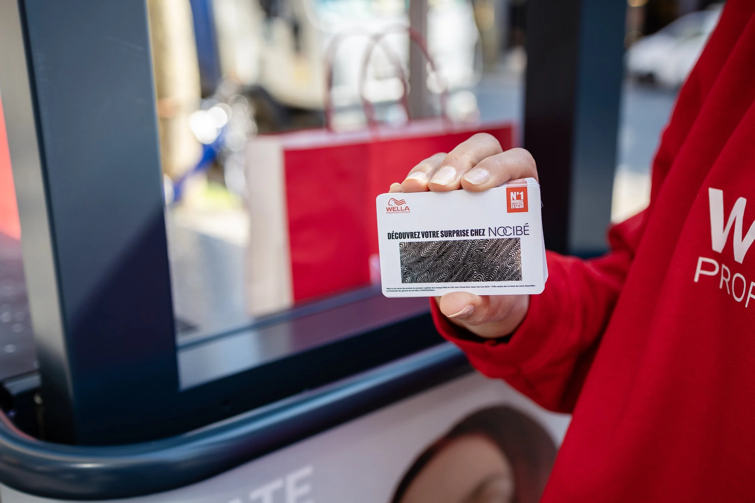 Une personne tient une carte de fidélité ou une carte promotionnelle devant une vitrine ou un comptoir, portant un sweat rouge velours avec le logo WELLA.
