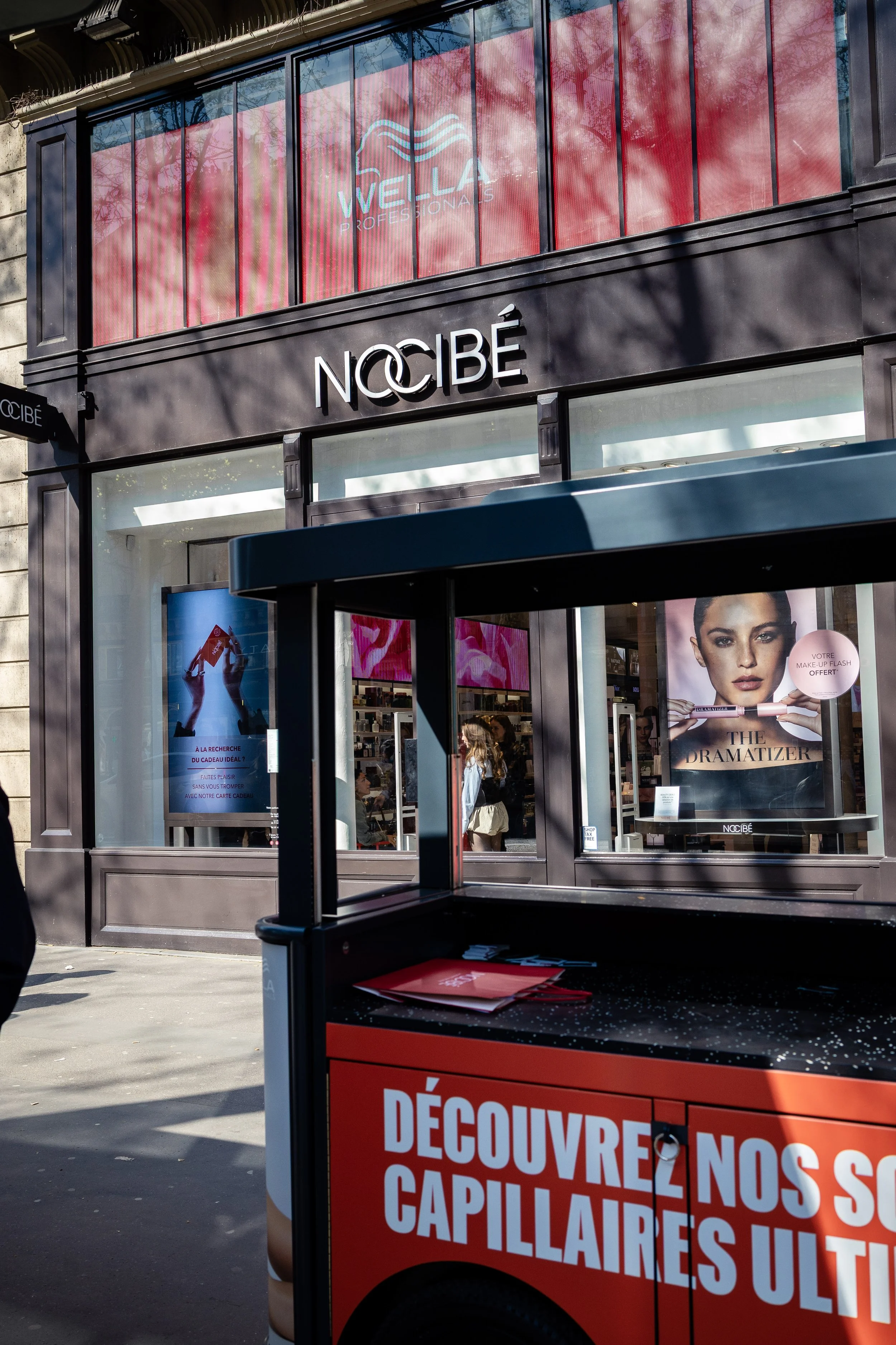 Façade d'un magasin Nocibé avec une publicité pour un produit de maquillage et un stand de promotion en premier plan affichant 'Découvrez nos nouveautés capillaires'.