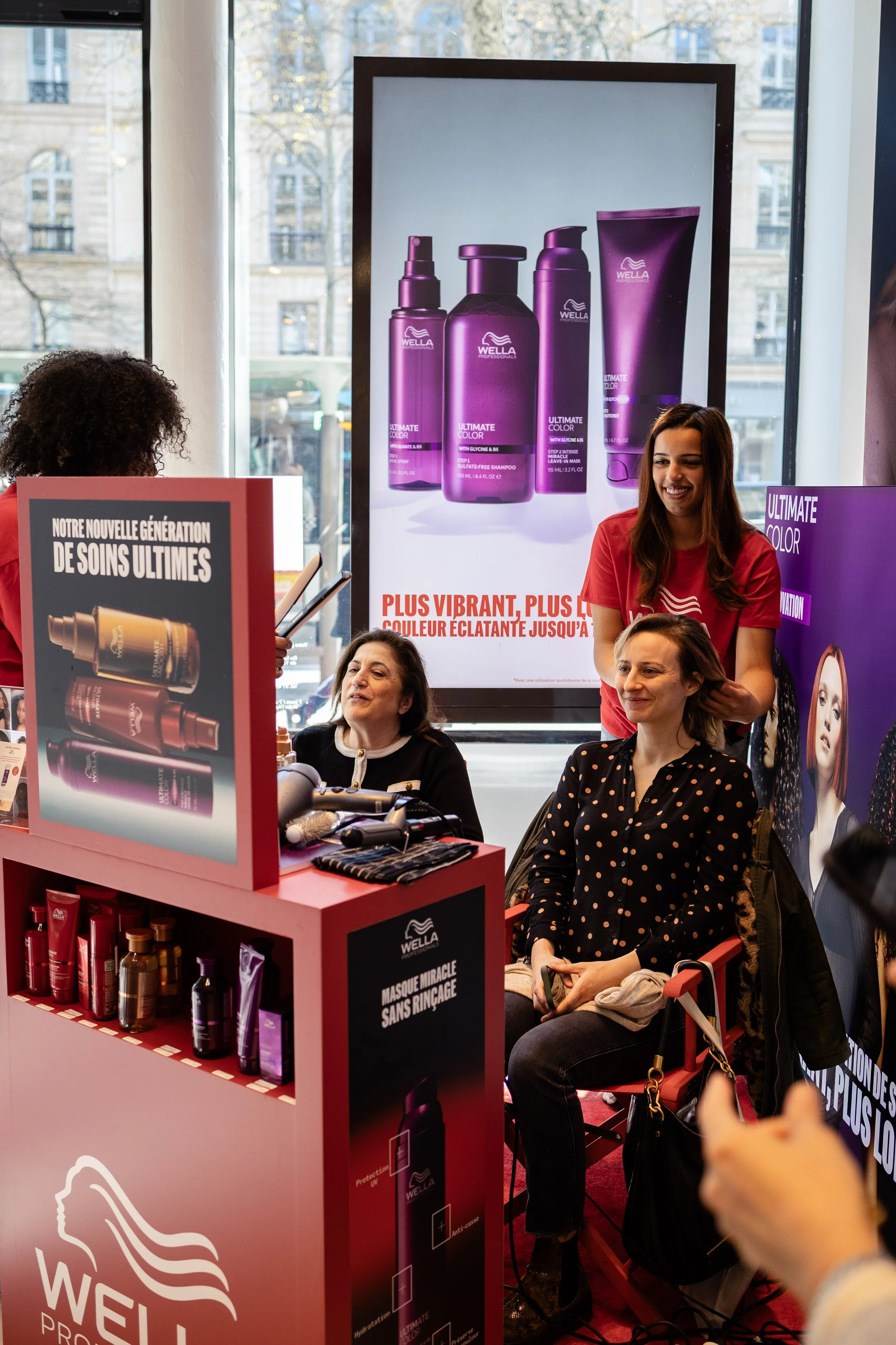 Une boutique de produits capillaires avec des affiches promotionnelles, des femmes discutant et des produits de la marque Wella.