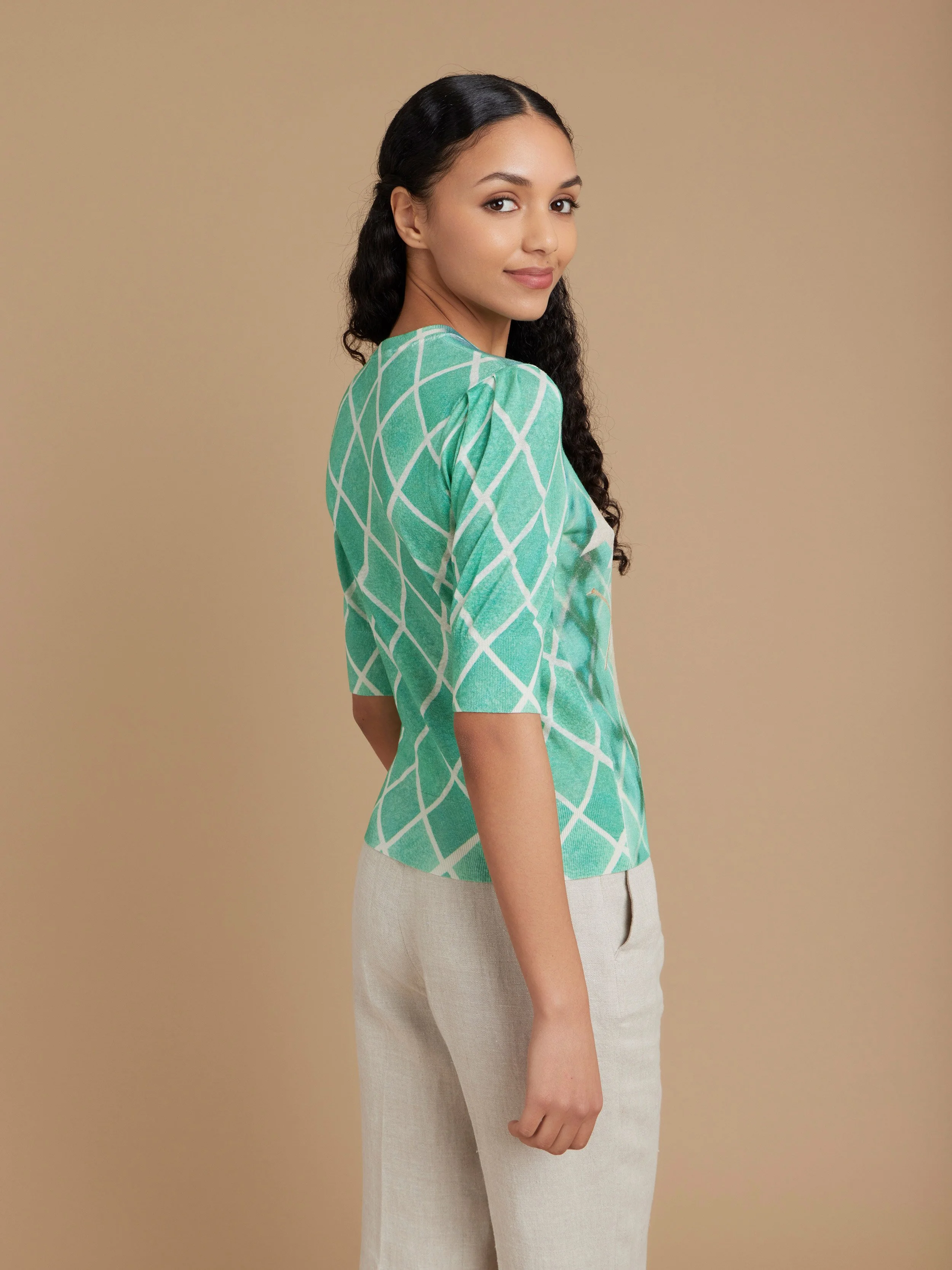 Une femme porte un haut vert à motifs géométriques et un pantalon beige, regardant par-dessus l'épaule avec un fond beige.
