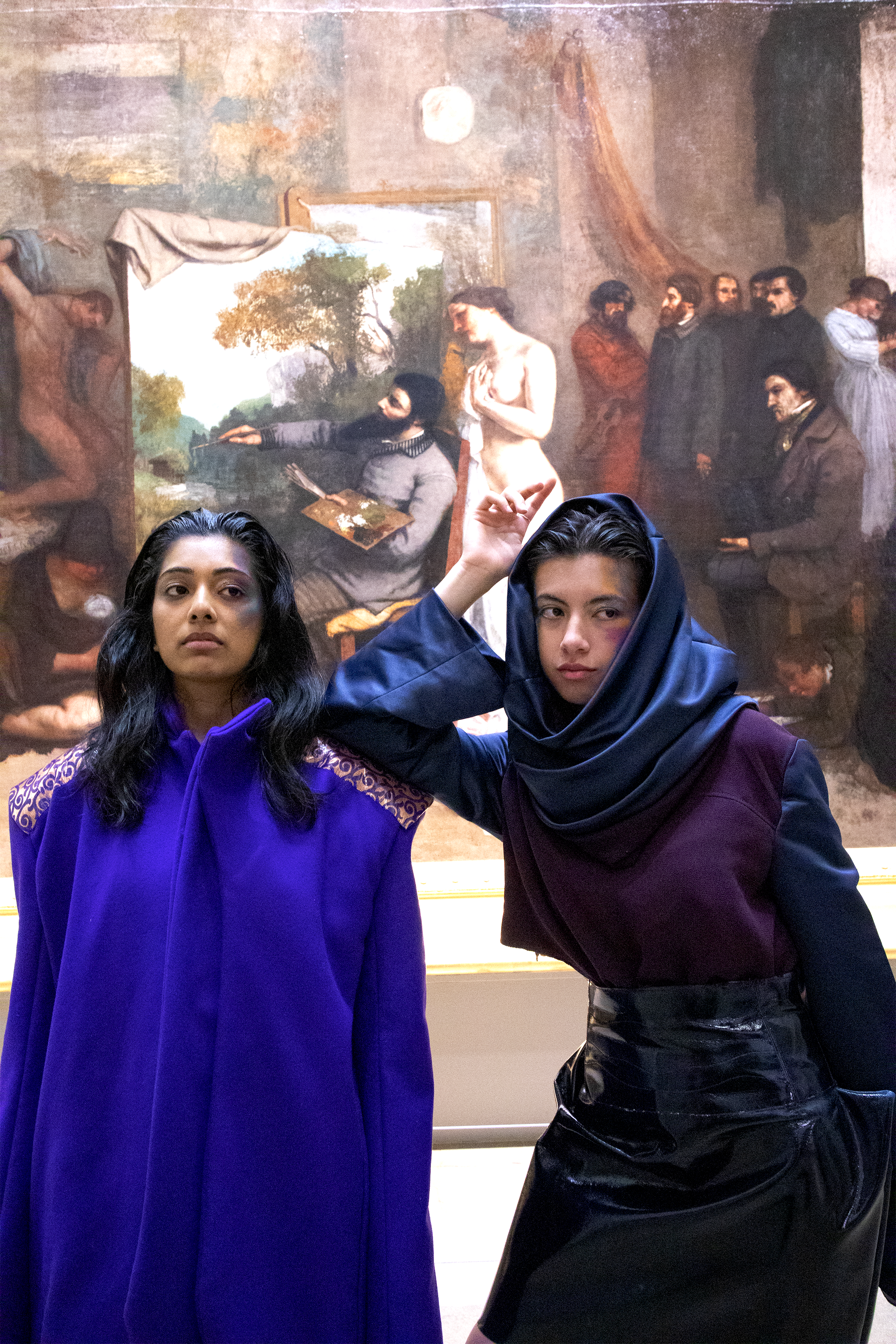 Deux femmes posent en face d'une peinture dans un musée. La peinture représente plusieurs personnes, dont une femme nue et une personne en train de peindre.