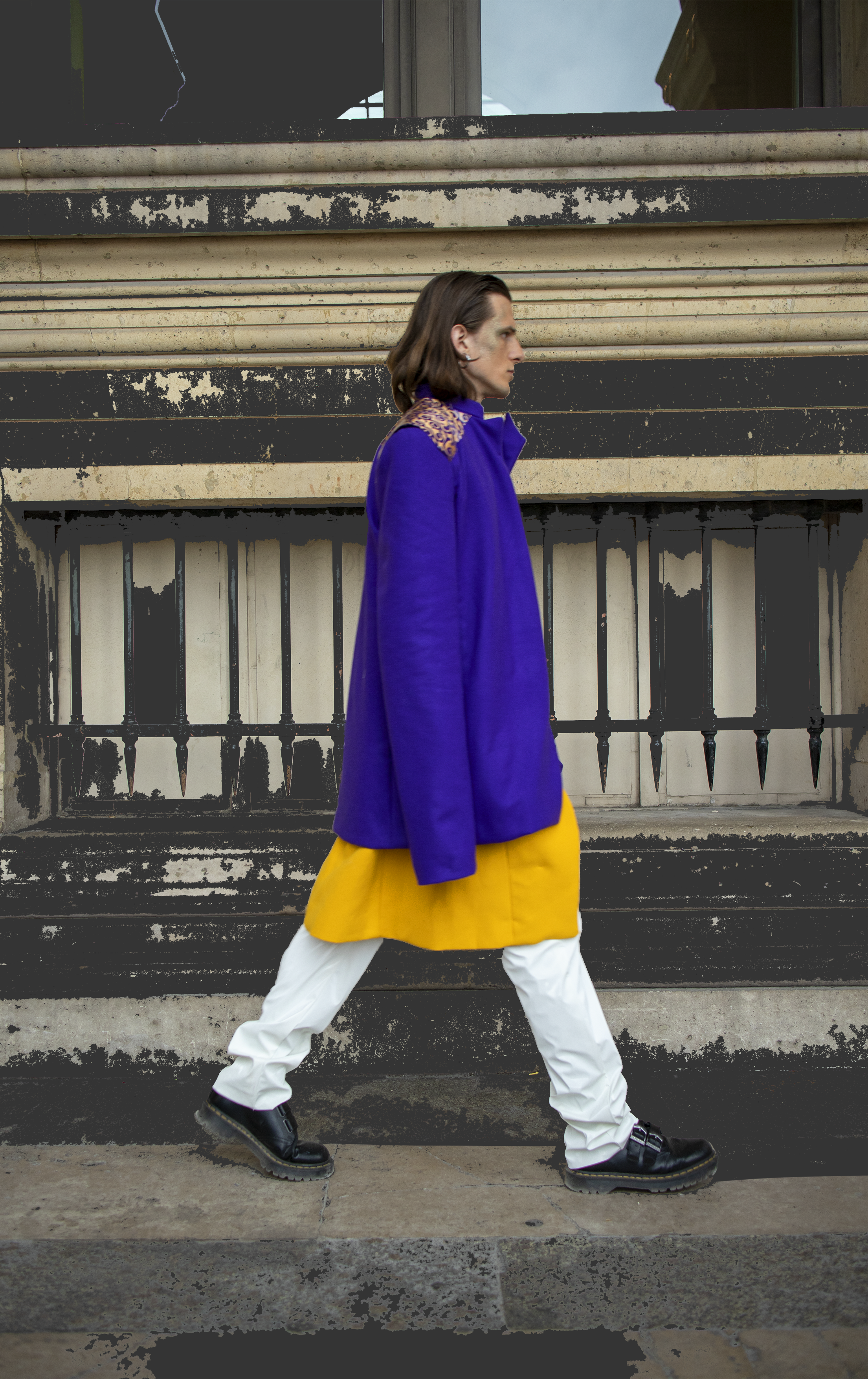 Un homme marche sur un trottoir, portant un manteau violet avec des épaulettes dorées, une jupe jaune, un pantalon blanc et des chaussures noires. En arrière-plan, un bâtiment urbain usé.