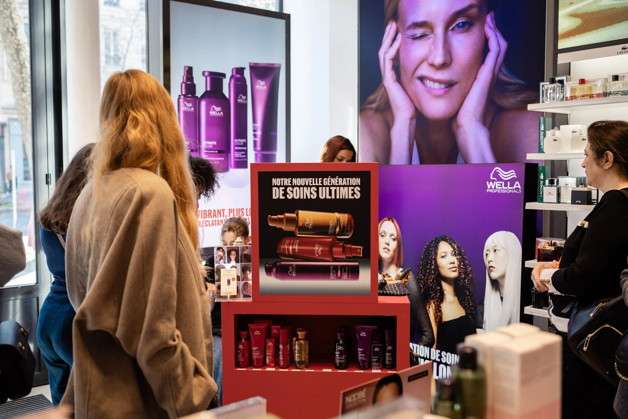Des personnes faisant la queue dans un stand de produits capillaires Wella dans un magasin. Il y a des affiches publicitaires et un présentoir avec des produits, notamment des shampooings et des sérums. Une grande image d'une femme souriante est visible à l'arrière-plan.