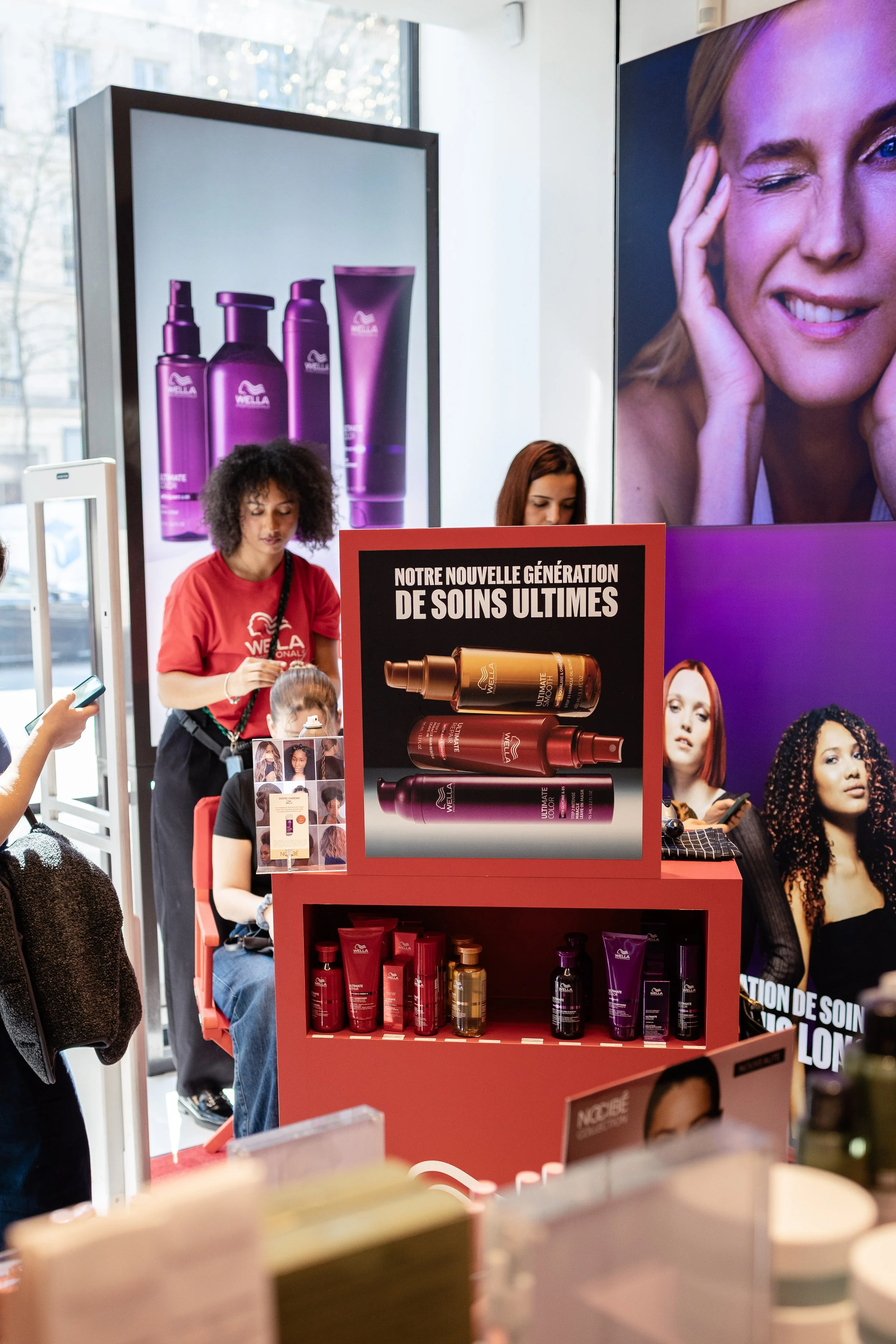 Stand de produits de soins capillaires avec affiches promotionnelles, plusieurs personnes utilisant leurs téléphones et un employé en rouge