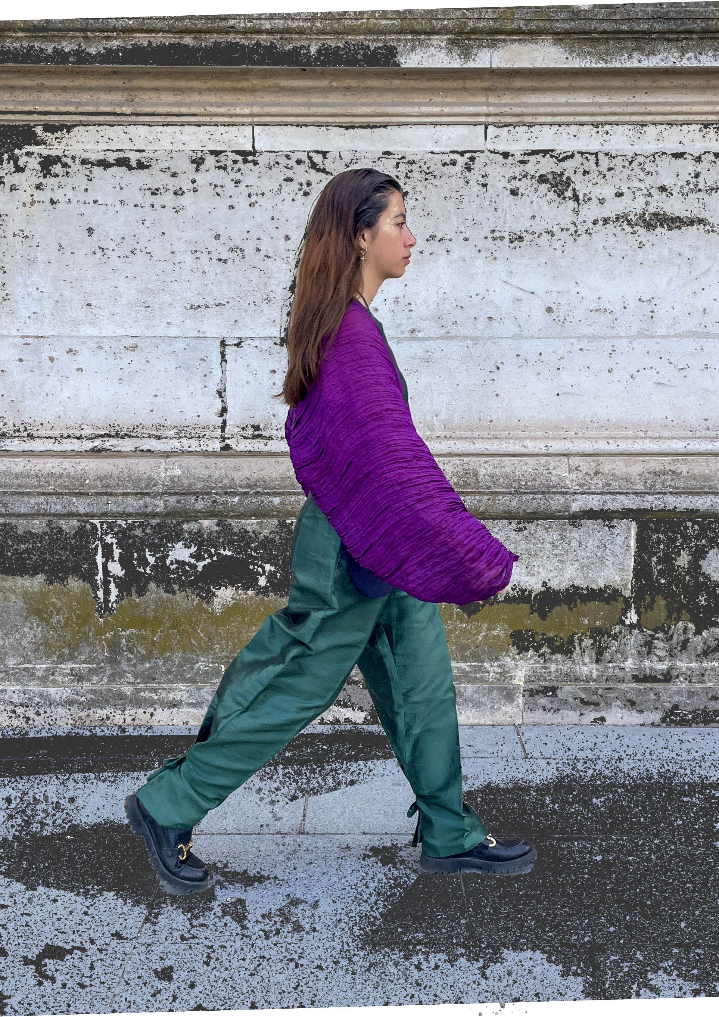 Femme marchant en passant devant un mur en pierre, portant une veste violette et un pantalon vert foncé, avec des chaussures noires.