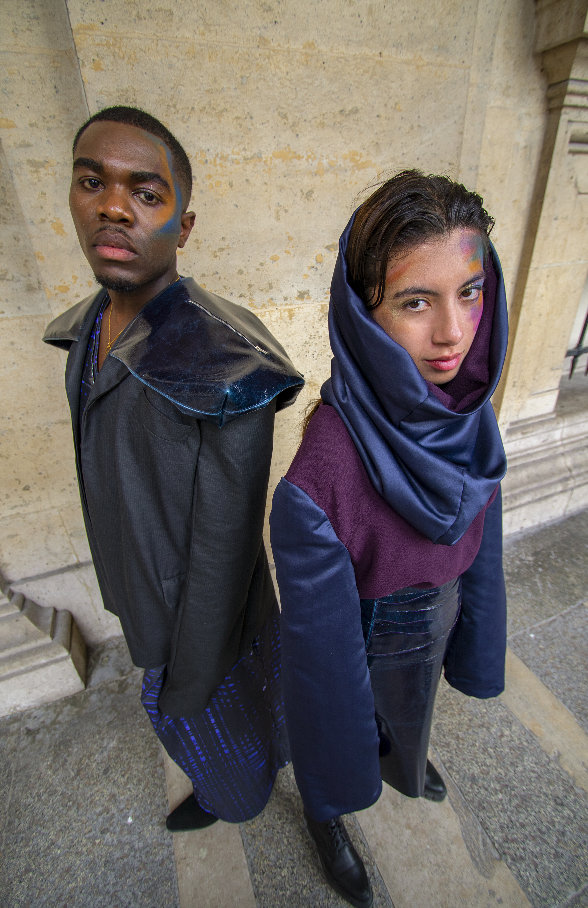 Deux jeunes adultes posent dos à dos contre un mur beige en pierre, portant des vêtements modernes avec des touches de couleur et du maquillage coloré sur leur visage.