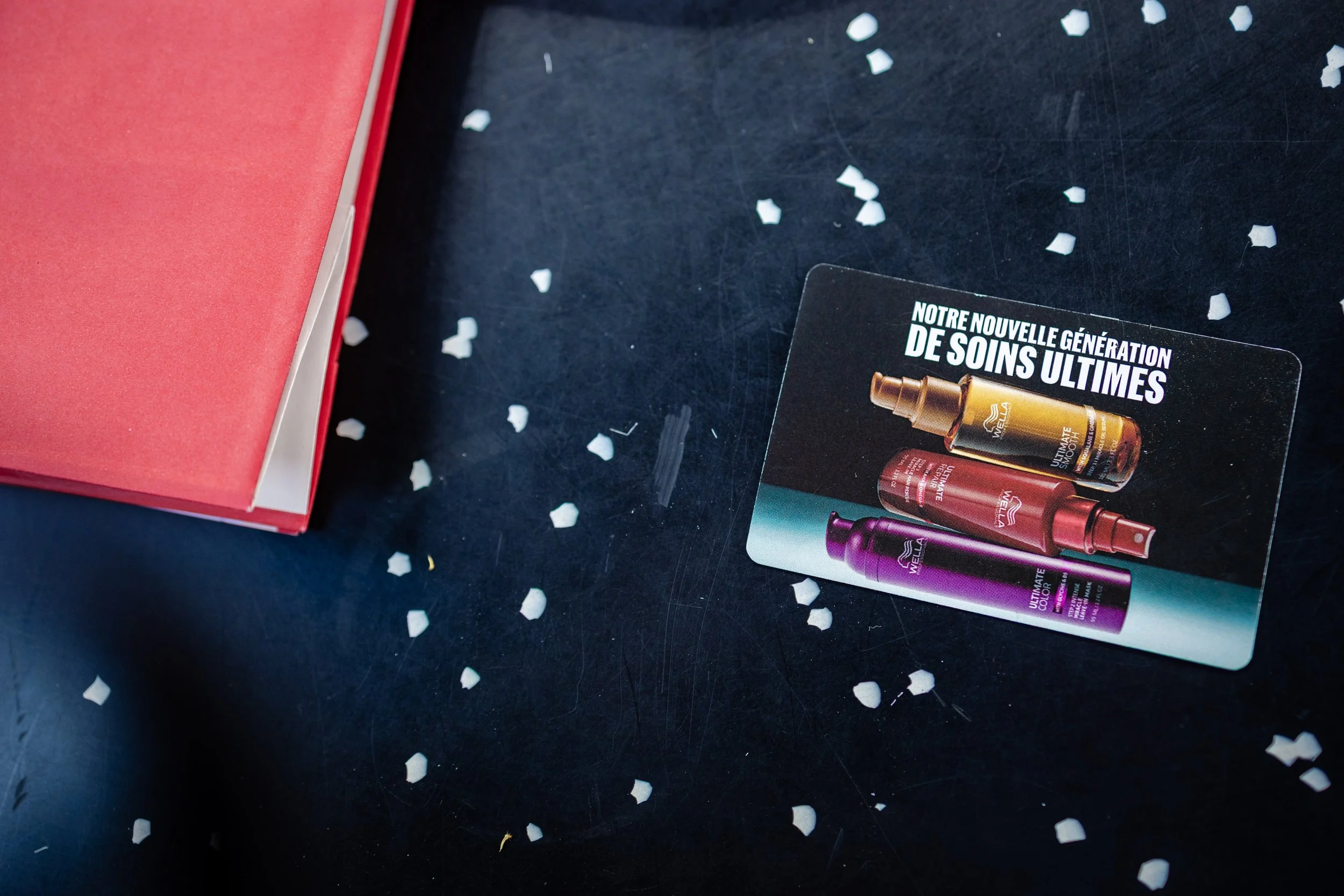 Une photo montrant une carte promotionnelle pour des soins ultimes avec trois petits flacons colorés (doré, rouge, violet), une boîte rouge dans le coin supérieur gauche, et des confettis blancs dispersés sur une surface noire.