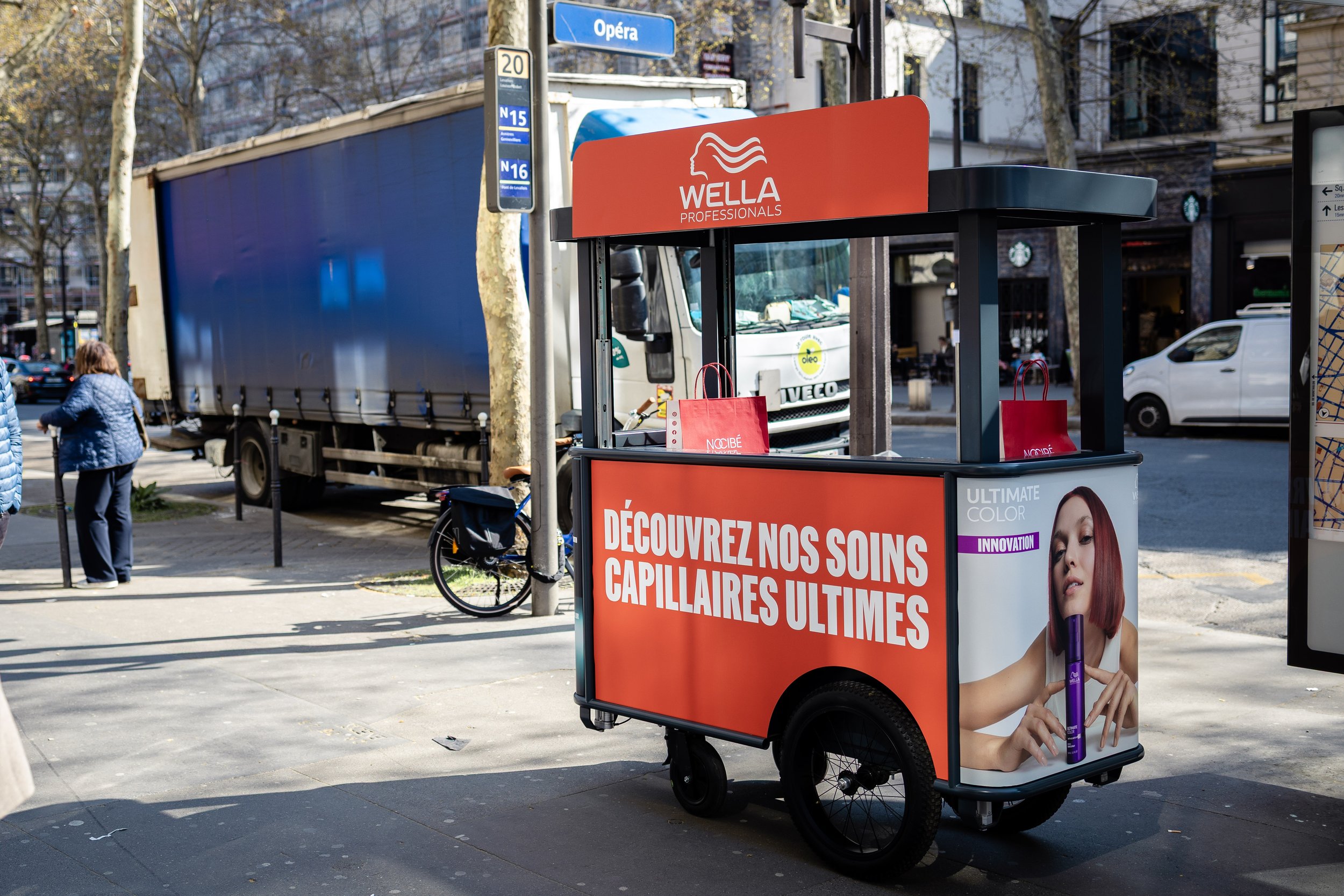 Chariot de vente de produits capillaires avec une affiche en français "Découvrez nos soins capillaires ultimes" et une image d'une femme avec les cheveux rouges, dans une rue urbaine.