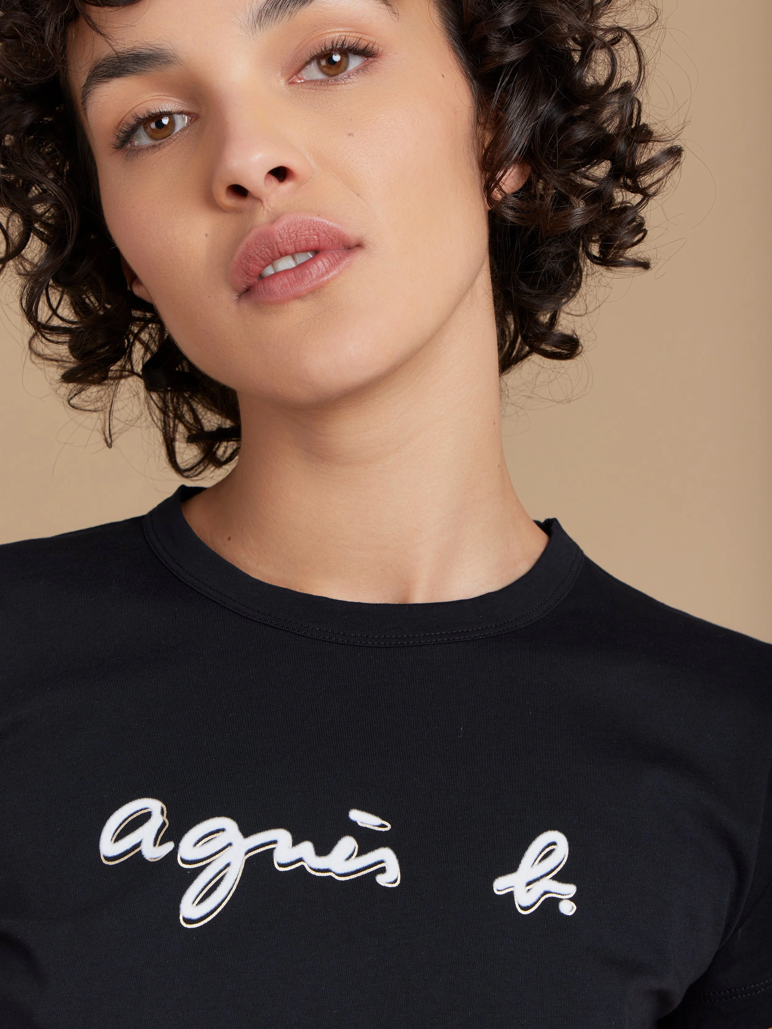 Jeune femme aux cheveux bouclés portant un t-shirt noir avec l'inscription "agüis t." en lettres blanches, pose devant un fond beige.