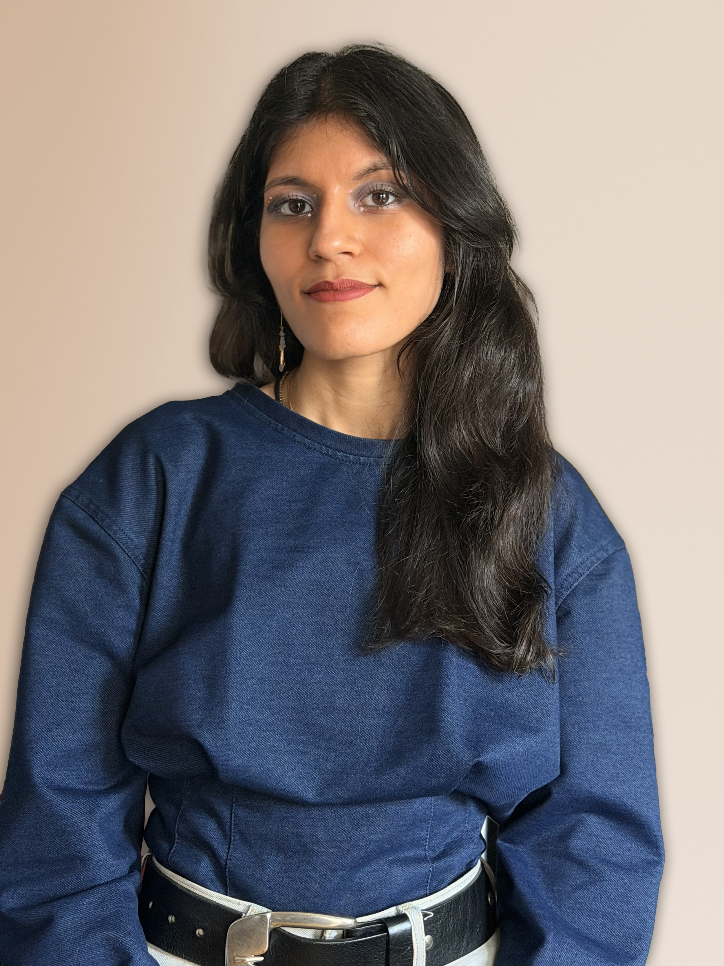 Jeune femme avec cheveux longs, vêtue d'un sweat bleu, portant une boucle d'oreille, se tenant devant un mur beige.